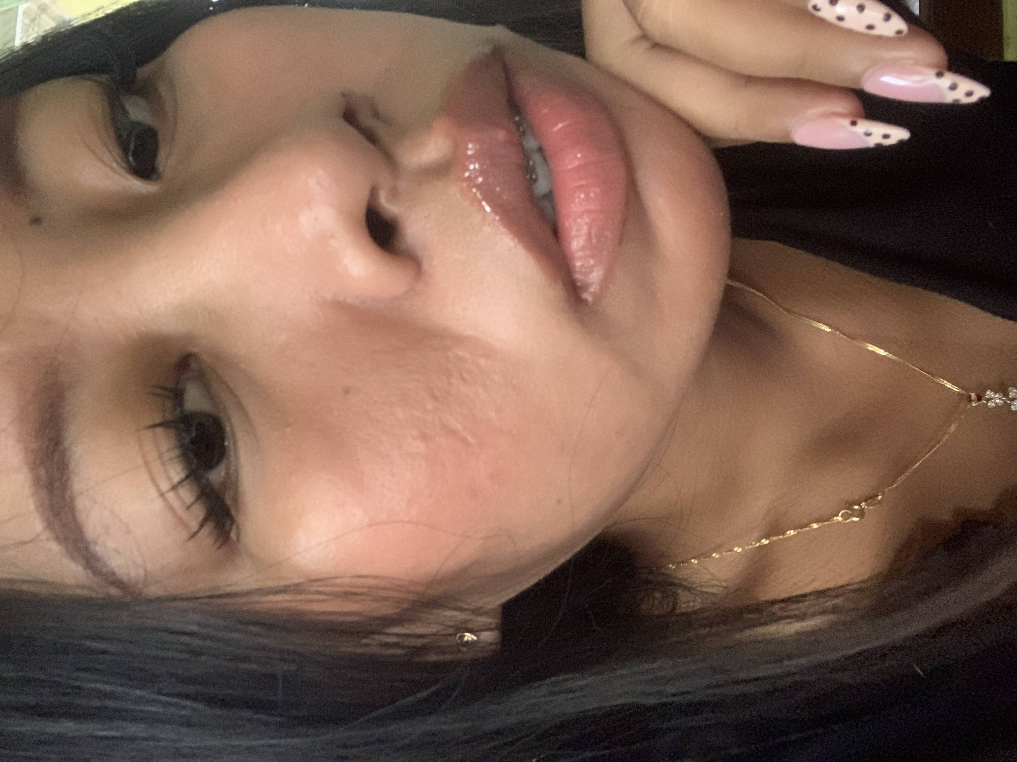 A close-up, low-angle, medium shot captures a young woman's face from the nose down, with her right hand supporting her chin. She has dark hair that falls around her face, and she is wearing a thin gold necklace with a small pendant. Her lips are glossy and a shade of pink, and her teeth are visible behind them, suggesting she might be smiling or speaking. She is also wearing braces. Her fingernails are painted pink with black polka dots in a French manicure style, and one nail is on her chin, while another is visible near her cheek. A small gold stud earring is visible on her left ear. The background is blurred, but it appears to be an indoor setting with some light and possibly furniture. There is no indication of the city of Jajag, Indonesia, or any specific location within it.