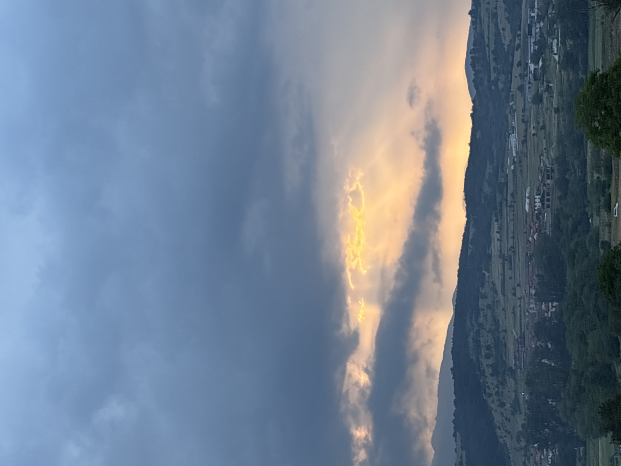 The image depicts a dramatic mountainous landscape under a dynamic sky, likely at sunrise or sunset in Strelcha, Bulgaria. Dark, heavy clouds dominate the left side of the frame, colored in shades of blue and gray. In the center, a striking burst of golden-yellow light pierces through a break in the clouds, creating a luminous, almost ethereal effect. These sun rays extend downwards and to the right, illuminating parts of the cloud cover and the distant landscape.
To the right, a range of rolling hills or mountains stretches across the image, their slopes appearing dark green and shadowed. Scattered across these hillsides are numerous buildings and small settlements, suggesting a rural or semi-rural community nestled within the terrain. Patches of lighter green indicate fields or pastures. The interplay of the dark clouds, intense golden light, and the shadowed mountains creates a high contrast scene, highlighting the beauty of natural light phenomena. The time of day is clearly either early morning or late evening, characterized by the warm, low-angle sunlight. There is no visible text in the image.