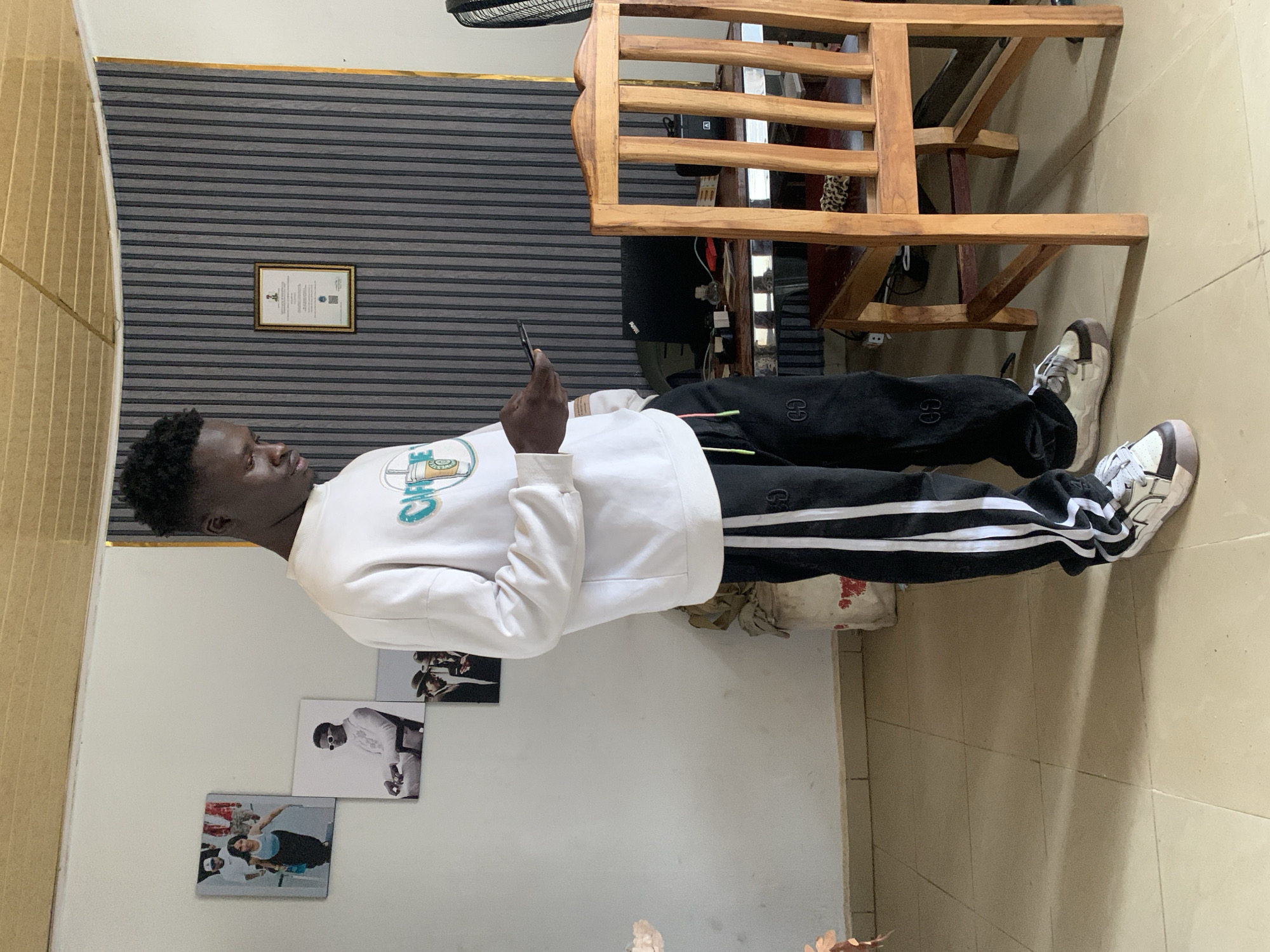 A young Black male stands in a room, holding a smartphone. He is wearing a white crewneck sweatshirt with a graphic design on the front and black track pants with white stripes. His hair is short and styled. The room has white walls, a wood-paneled ceiling, and tiled flooring. On the wall to the left, there are three framed pictures. To the right, a wooden chair sits at a desk with a laptop and other items. The background features a wall covered in vertical grey and white striped wallpaper. The image appears to be taken indoors, possibly in a home or office setting. The location is identified as Gura Top, Nigeria.