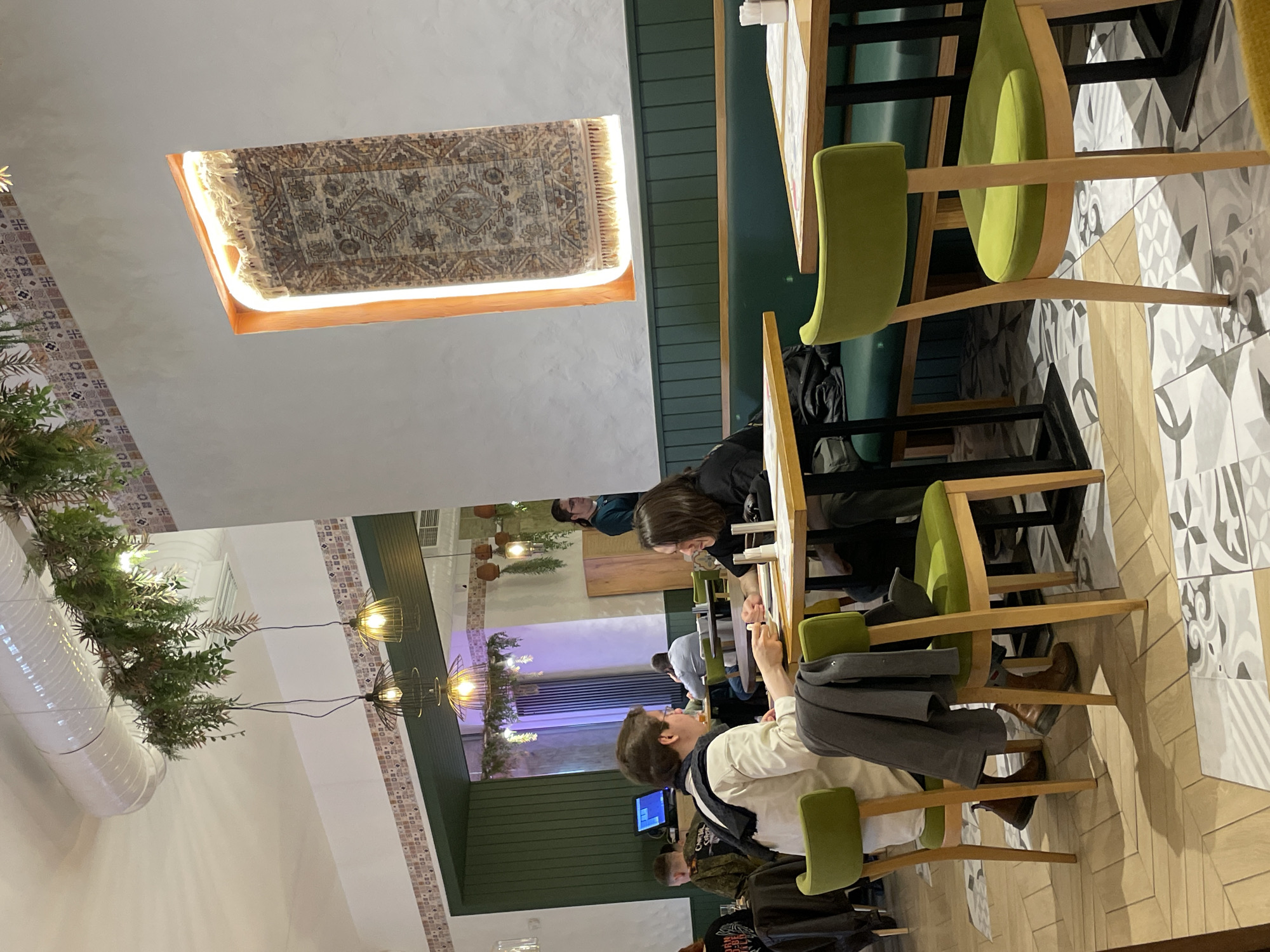 The image shows the interior of a restaurant. People are seated at tables. The decor features green accents, light-colored walls, and tiled flooring. An ornate rug is displayed as a decorative element on the ceiling with a light fixture on its edges. Hanging light fixtures and artificial greenery are also present. The location is most likely Saint Petersburg, Russia.