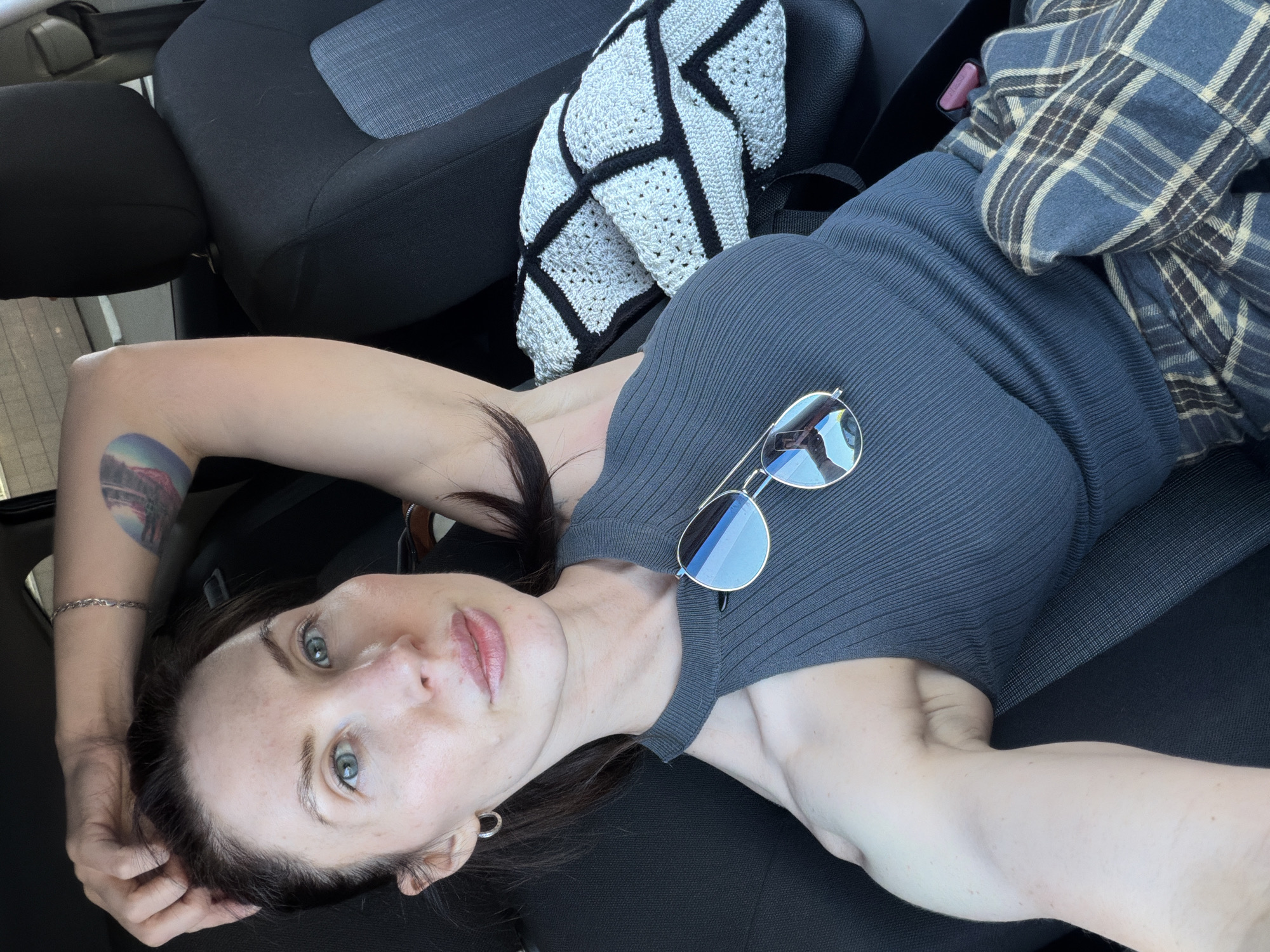 A young woman lies in the passenger seat of a car. She wears a gray ribbed top and has sunglasses resting on her chest. She has a tattoo on her left arm. A black and white crocheted blanket and a blue plaid shirt are nearby. The car's interior is black. There is no visual information to indicate that the location is in the city of Perm, Russia.