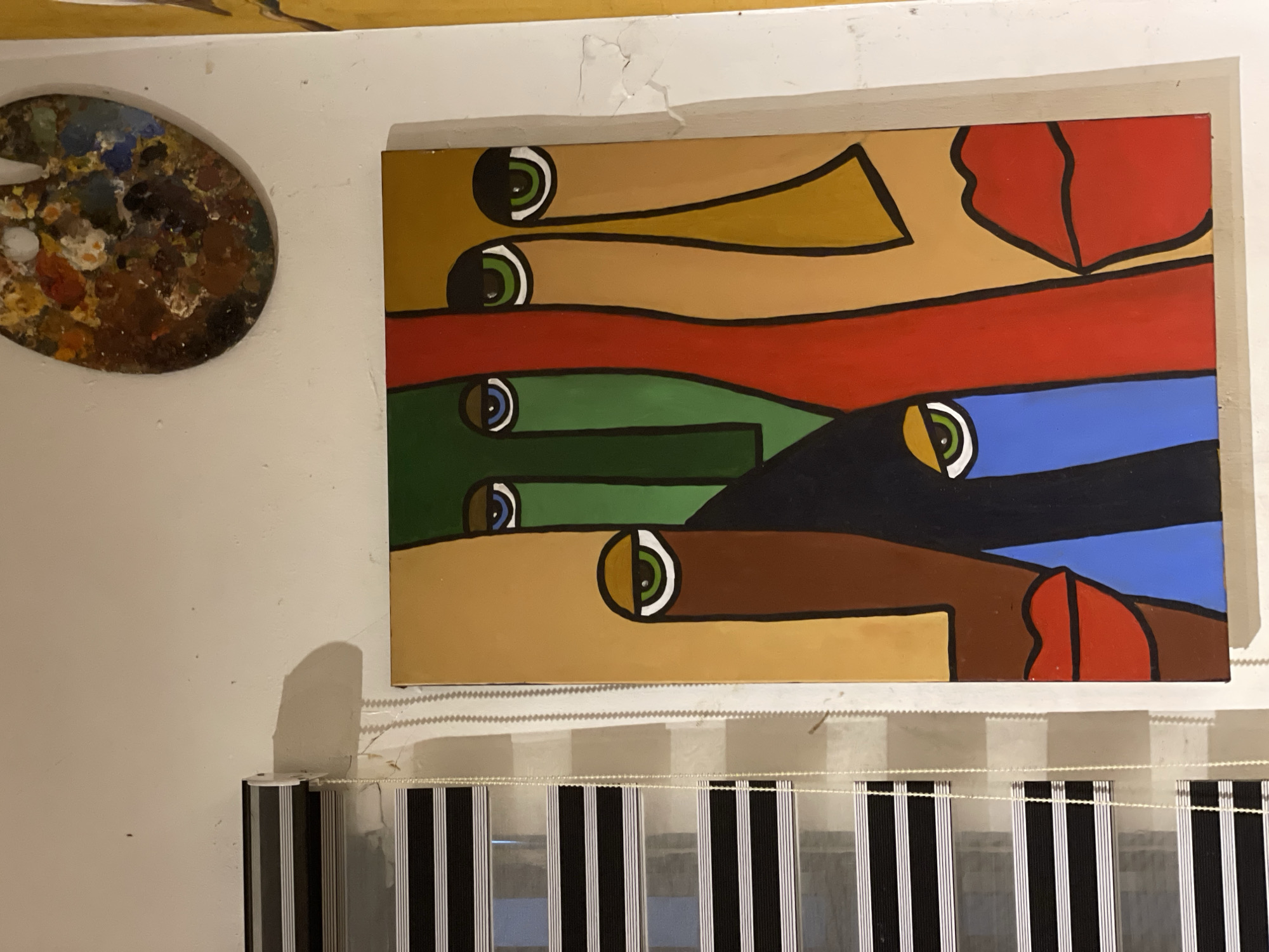The image depicts a wall in a dimly lit indoor setting, likely an art studio or gallery. A framed, abstract painting featuring several stylized, cubist-inspired faces with bold colors dominates the center of the frame. To the left of the painting are partially visible window blinds with black and white stripes. Above and to the right of the painting, a wooden artist's palette splattered with various colors of paint is mounted on the wall. The wall itself is white and shows signs of minor cracking and staining. The overall impression is that of an artistic space, possibly in Ado, Nigeria, given the context provided.