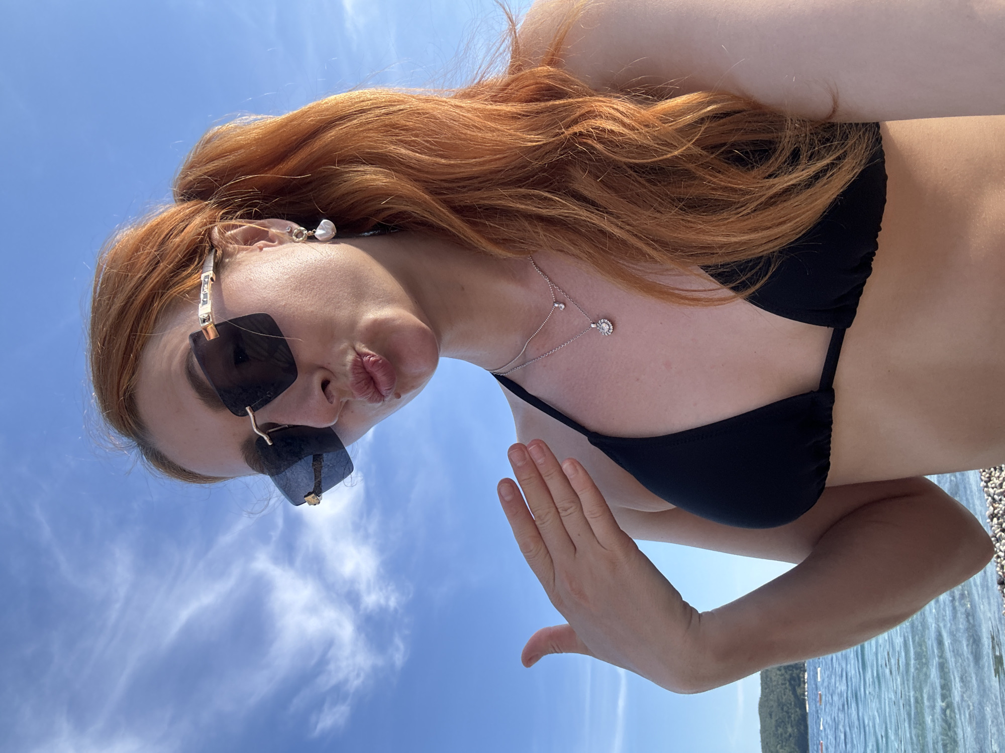A woman is taking a selfie on the beach in Budva, Montenegro. She is wearing a black bikini and sunglasses, and has reddish-orange hair. She is pouting and holding her hand up in front of her face. The sky is blue with some clouds, and the water is visible in the background. The beach appears to be pebbled.