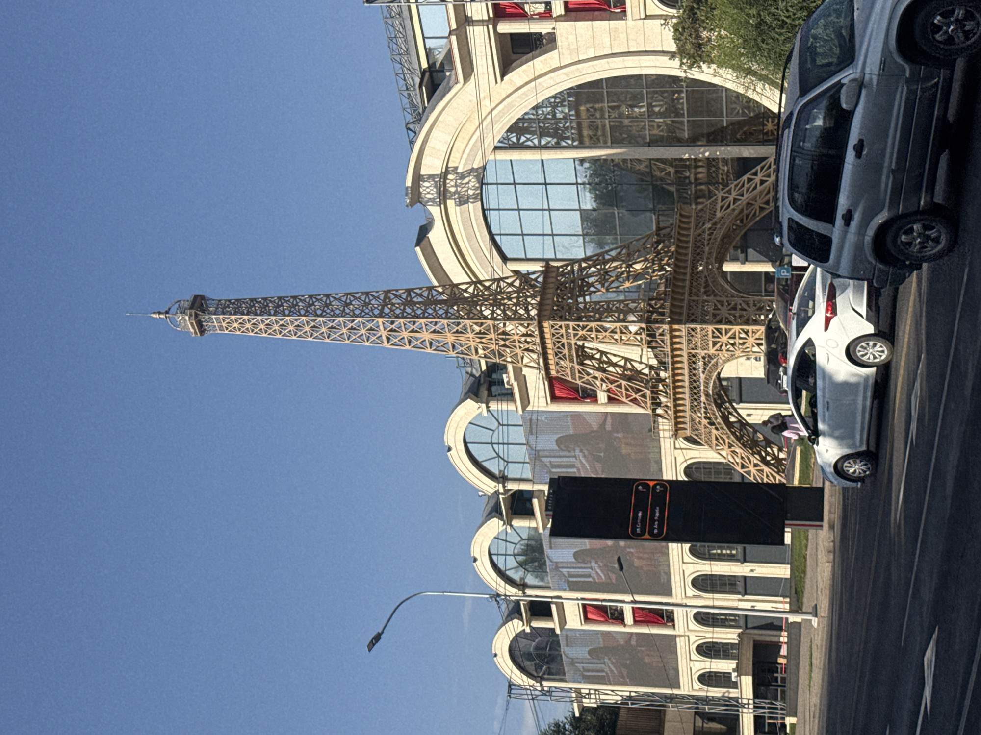 The image shows a daylight view of a street scene in Almaty, Kazakhstan, featuring a replica of the Eiffel Tower as part of the facade of a building. The Eiffel Tower replica is made of wood and is positioned in front of a large, cream-colored building with arched windows and large glass panels. Red curtains are visible behind some of the windows. A digital billboard is located to the left of the Eiffel Tower replica. On the street in front of the building, two cars are visible: a white sedan and a silver SUV. The sky is clear and blue. The scene appears to be a commercial area.