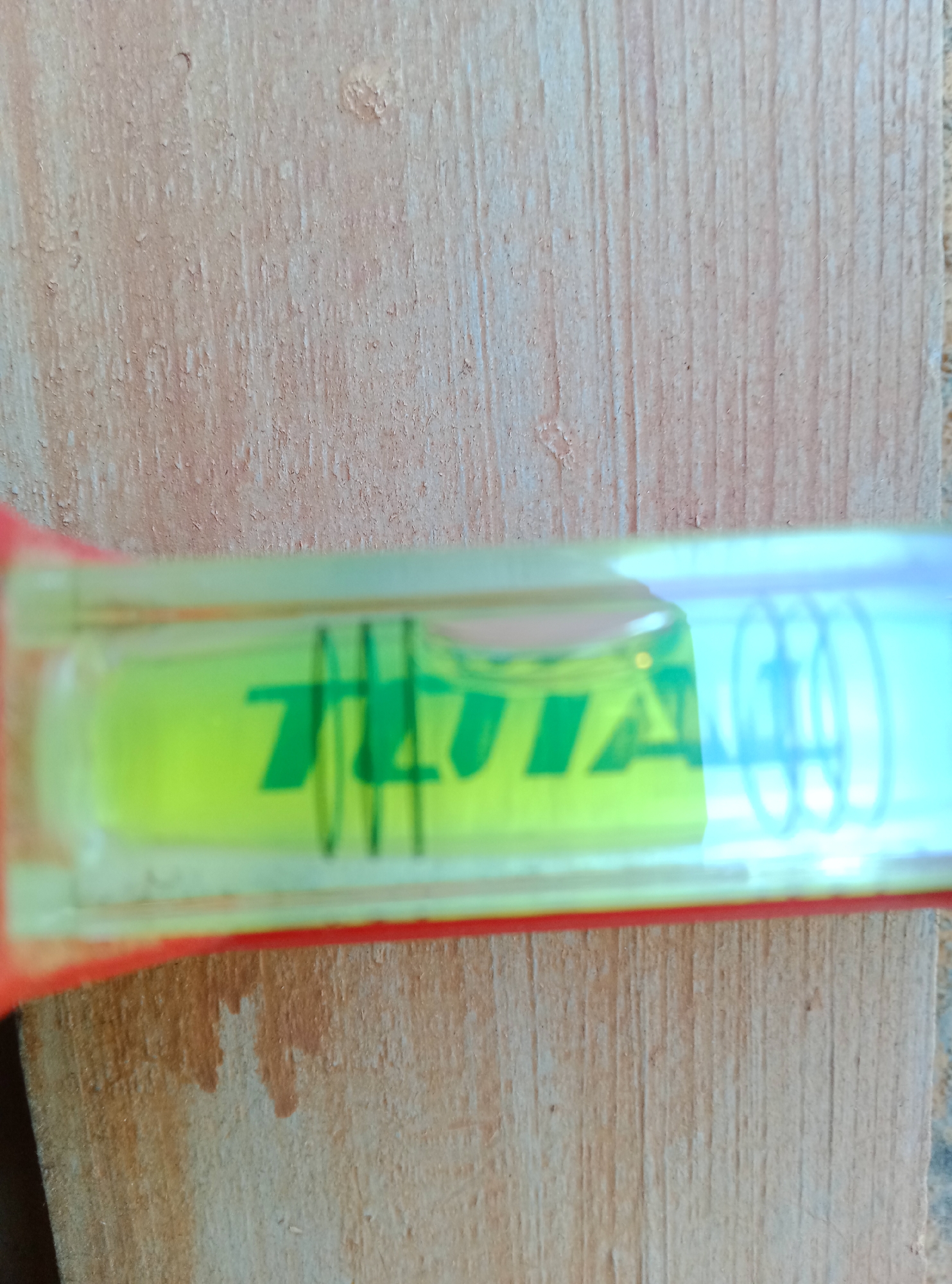 The image is a close-up, horizontal shot of a spirit level resting on a wooden surface. The spirit level is a transparent tube filled with bright yellow liquid and contains a single air bubble. Green markings, possibly indicating a measurement or brand name, are visible within the liquid. The wooden surface appears to be unfinished and has a visible grain pattern. The background shows more of the wooden surface. The image does not provide any information about the city of Pshap, Abkhazia, or any human presence.