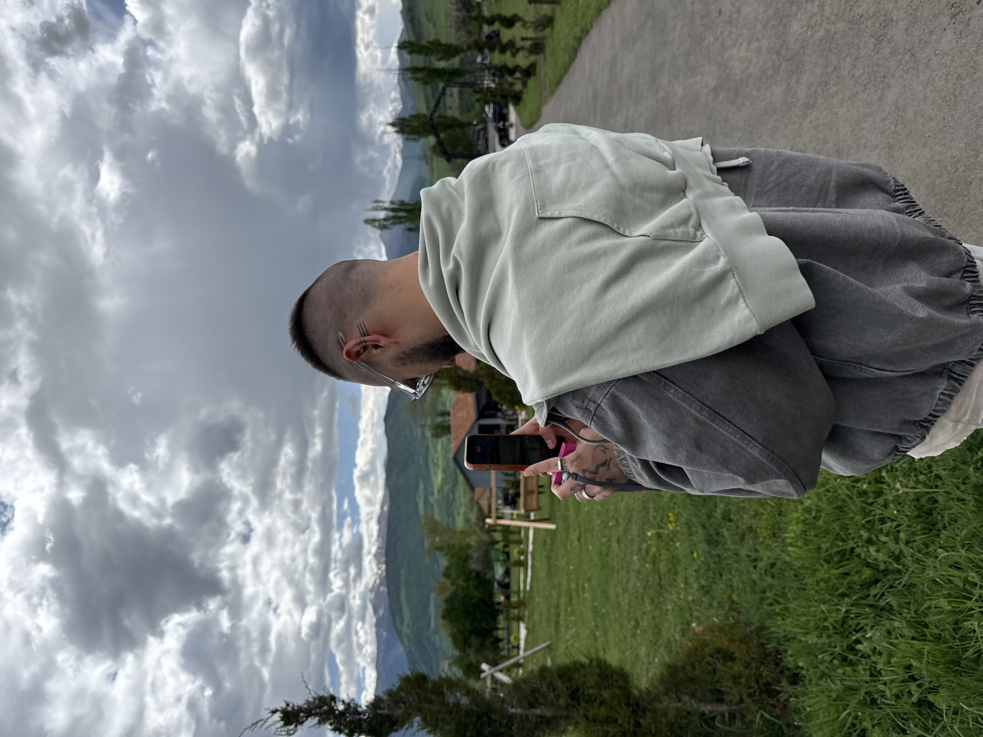 A man is standing outdoors, holding and looking at a smartphone. He has short hair and is wearing glasses. He's wearing a light-colored hooded sweatshirt over a darker grey jacket. His hands and arms are tattooed, and he's wearing rings. The background shows a green field, mountains, and a cloudy sky. The location appears to be Dsegh, Armenia, based on the presence of mountainous terrain and greenery common to the region.