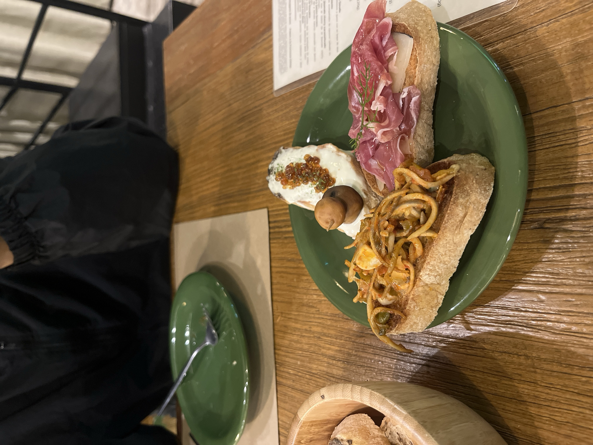 A green plate sits on a wooden table in a restaurant in San Andrés Cholula, Mexico. The plate contains three open-faced sandwiches or tapas. One is topped with what appears to be prosciutto, cheese, and dill. Another has a white topping with orange roe and two small sausages. The third is covered with noodles. Next to the green plate is another empty green plate with a fork on it. A bamboo container with bread sits at the bottom of the shot. A person wearing a black jacket is in the left side of the image.