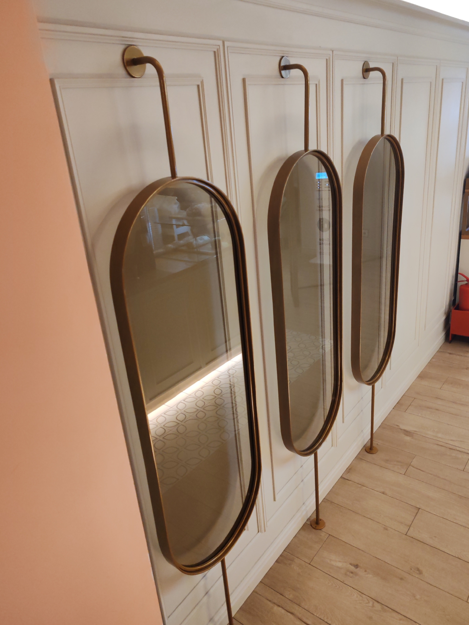 This interior image features three tall, narrow oval-shaped mirrors, each with a brass-toned frame and slender brass supports. The mirrors are mounted against a light cream or off-white wall adorned with classic rectangular paneling. Each mirror is suspended from the top by a curved brass rod that attaches to the wall, and further stabilized by a thinner brass rod extending downwards to the floor. To the far left, a smooth, unpaneled wall or partition in a soft peachy-pink hue adds a contrasting color element.
The floor of the visible space is covered with light-toned wooden planks. However, reflections in the mirrors reveal a different floor type further into the room or in an adjacent area, featuring light-colored tiles with a subtle, repeating geometric pattern. The reflections also show indistinct elements of the surrounding interior, including overhead lighting and what appears to be a red fire extinguisher cabinet in the reflection of the rightmost mirror. Some blurred, light-colored text or numbers, possibly including a 'b' or '0', are faintly visible in the reflection of the central mirror. The scene depicts a well-lit, modern yet elegant interior space, likely a commercial or public establishment such as a hotel, cafe, or restaurant, located in Saint Petersburg, Russia. No human activity is depicted.
