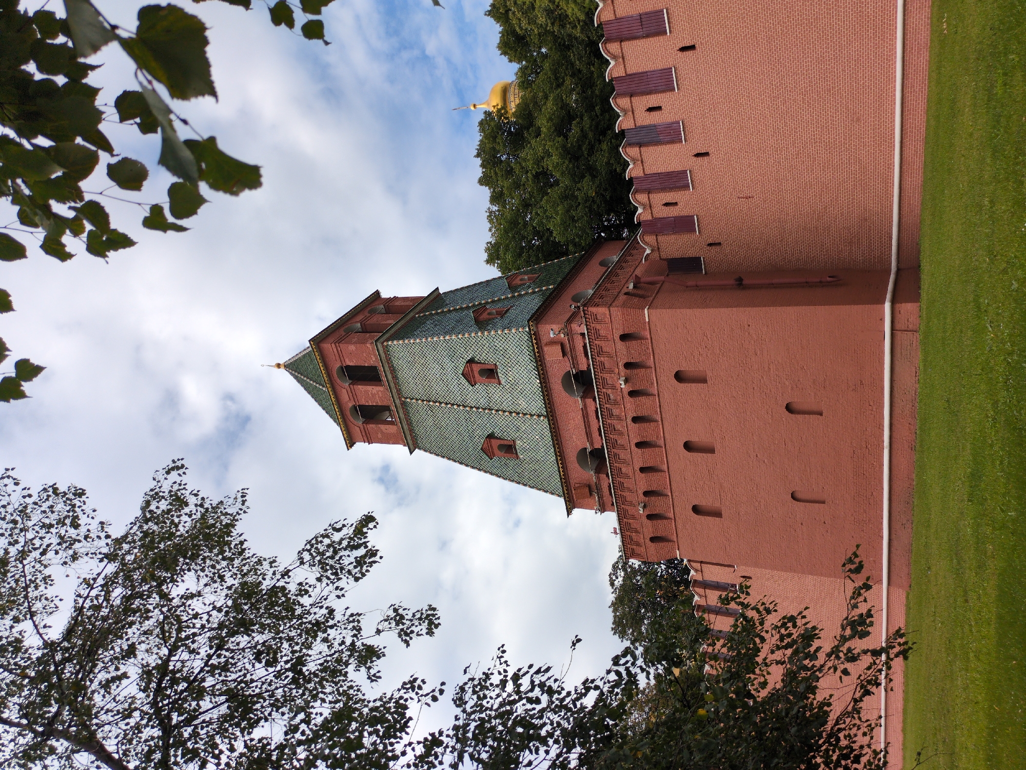 The image shows a tall, red brick tower with a green, faceted roof, part of a larger defensive wall. The tower has arched openings and small, rectangular windows. The wall is made of red brick and has crenellations along its top. Trees with green foliage are visible on the right side of the image, and a golden dome is peeking out from behind them. The foreground consists of a grassy lawn. The sky is cloudy and blue. The scene is set in a place that appears to be of historical significance, likely a fortress or a palace complex.