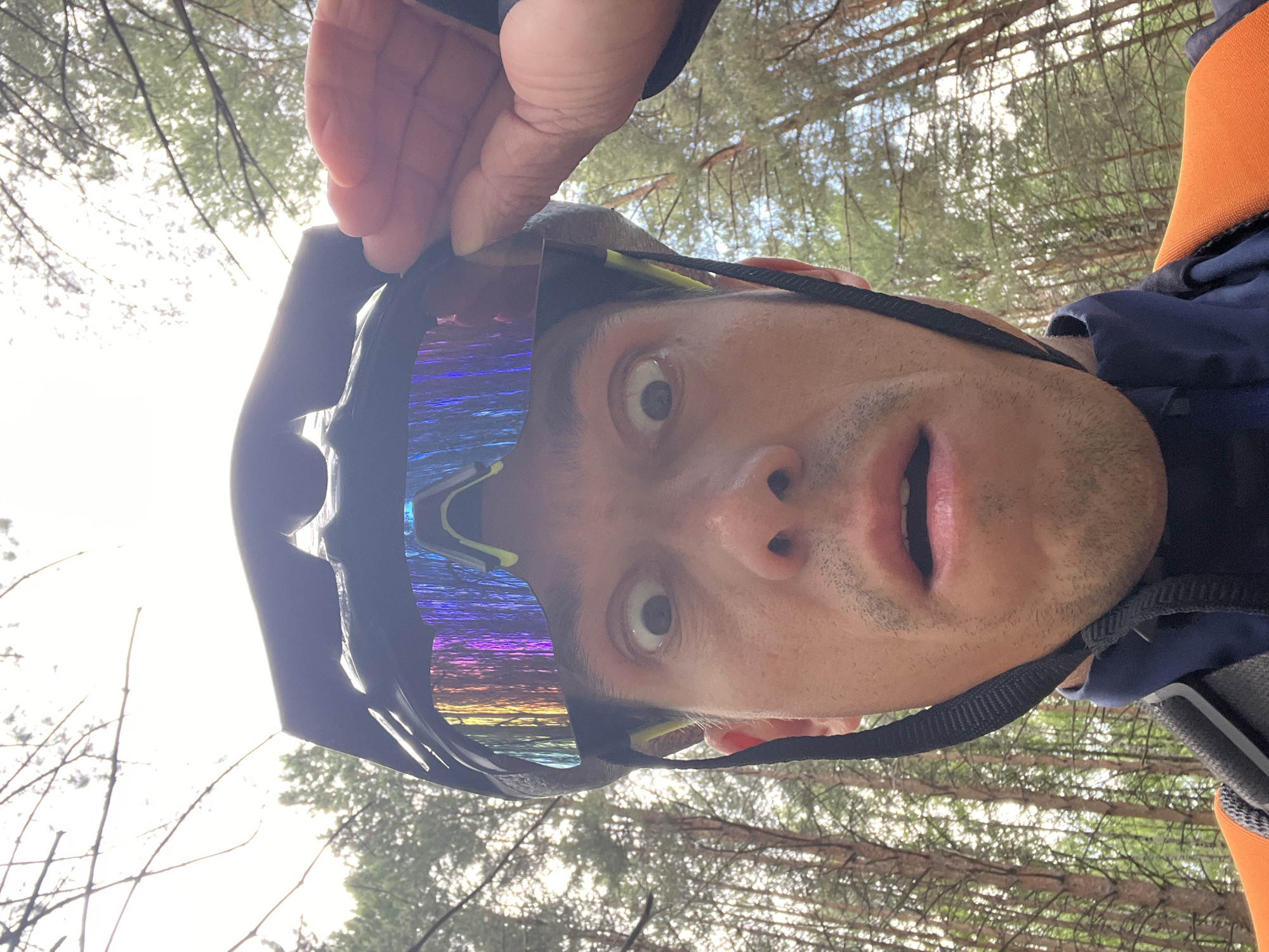 A man is taking a selfie in a forest. He is wearing a black bicycle helmet and iridescent cycling glasses. His right hand is raised to adjust or touch his sunglasses. He has a surprised expression on his face, with wide eyes and an open mouth. He is wearing a dark blue jacket and an orange vest. The background consists of tall trees with green foliage, suggesting he is in a wooded area. The lighting is bright, indicating it is daytime. There are no discernable urban elements or indications of the city of Chaikovsky, Russia.