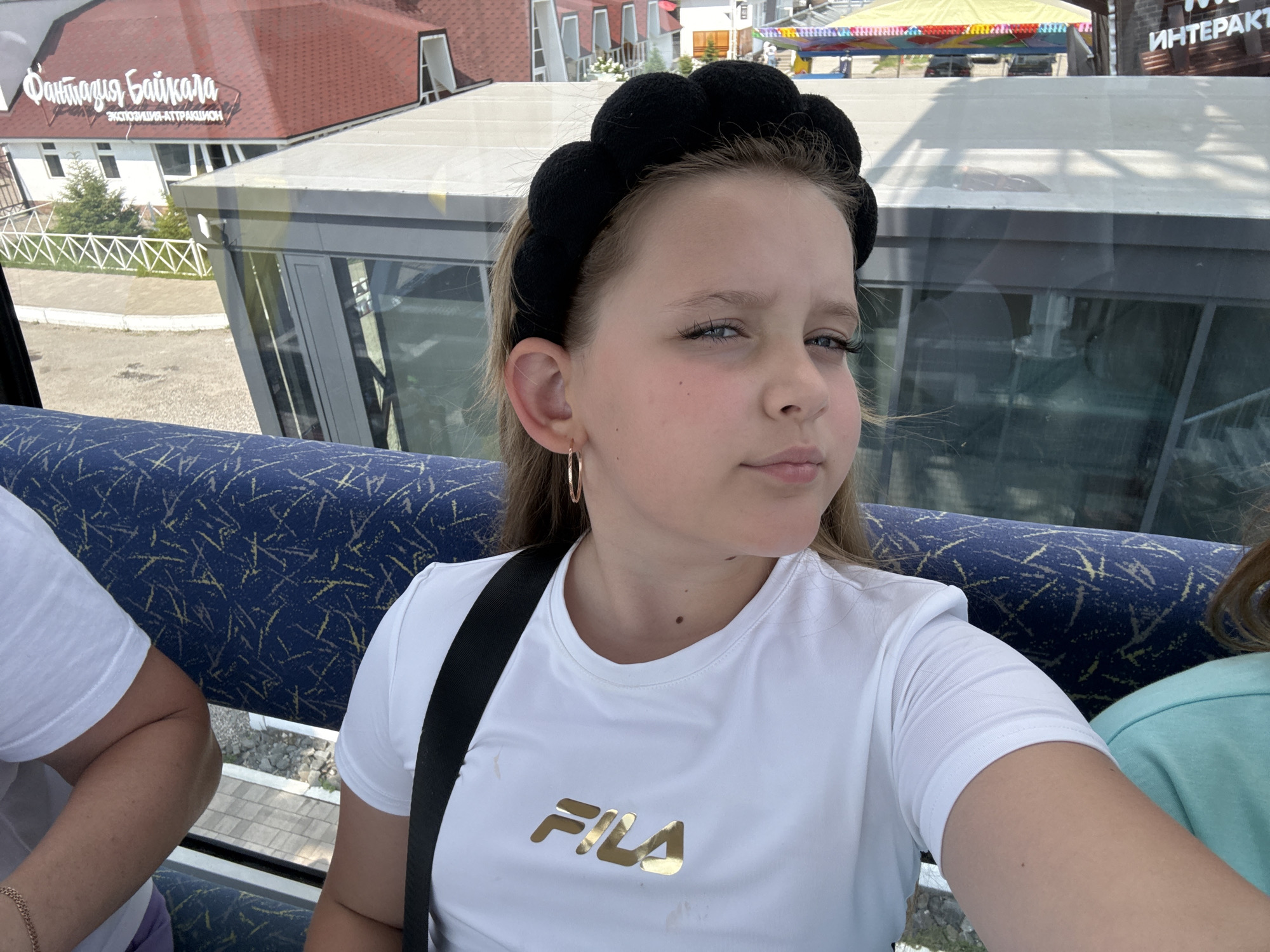 A young girl with blonde hair, wearing a black plush headband, a white t-shirt with a gold FILA logo, and gold hoop earrings, is looking directly at the camera. She is seated inside what appears to be a cable car or a funicular, with blue and yellow patterned seating visible behind her. The girl is in the foreground, with the upper body of another person on her left, also wearing a white t-shirt. Through the window of the cable car, buildings with red roofs and the text "Фантазия Байкала" (Fantasy of Baikal) are visible, indicating the location is likely Listvyanka, Russia, on the shore of Lake Baikal. The background also shows some colorful tents and structures, possibly part of an amusement park or tourist attraction. The overall scene suggests a sunny day and a tourist activity.