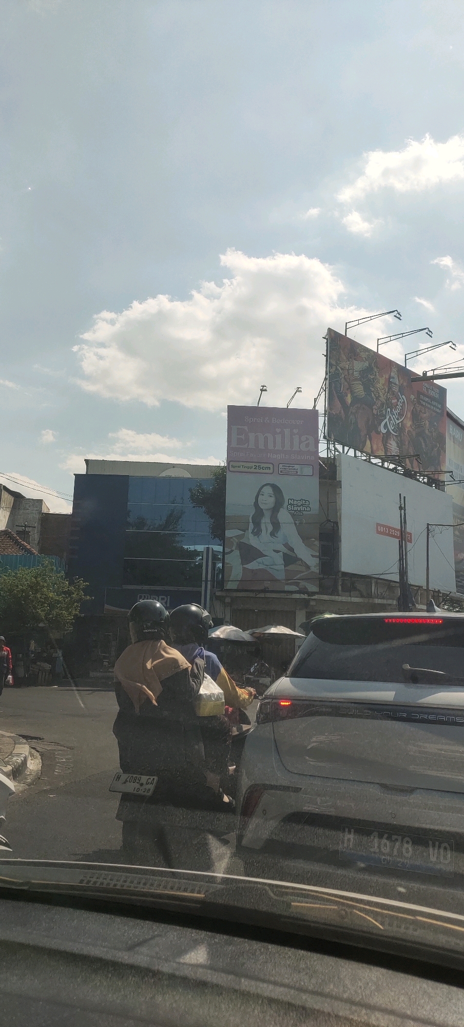 In Semarang, Indonesia, on a cloudy day, a traffic scene unfolds on a street. The foreground is dominated by the interior of a vehicle, likely a car, with its windshield and dashboard visible. Beyond the vehicle, a motorcycle carrying two individuals in helmets and protective gear is stopped in traffic. The driver, wearing a brown hijab, faces forward, while the passenger behind appears to be holding a yellow food container.
Further back, a white SUV with its taillights illuminated is also stationary. A license plate on this SUV reads "H 1678 VO". To the left, a building with a dark blue facade and reflective windows stands.
Dominating the background are several large billboards. One billboard, featuring an advertisement for "Emilia" bed covers and showcasing a woman, is prominently displayed. Next to it, a larger billboard displays a dynamic scene with figures, possibly from a movie or event advertisement. The sky above is a mixture of light blue and scattered clouds, suggesting daylight hours. The street appears to be paved, and the overall atmosphere suggests a typical urban traffic condition.