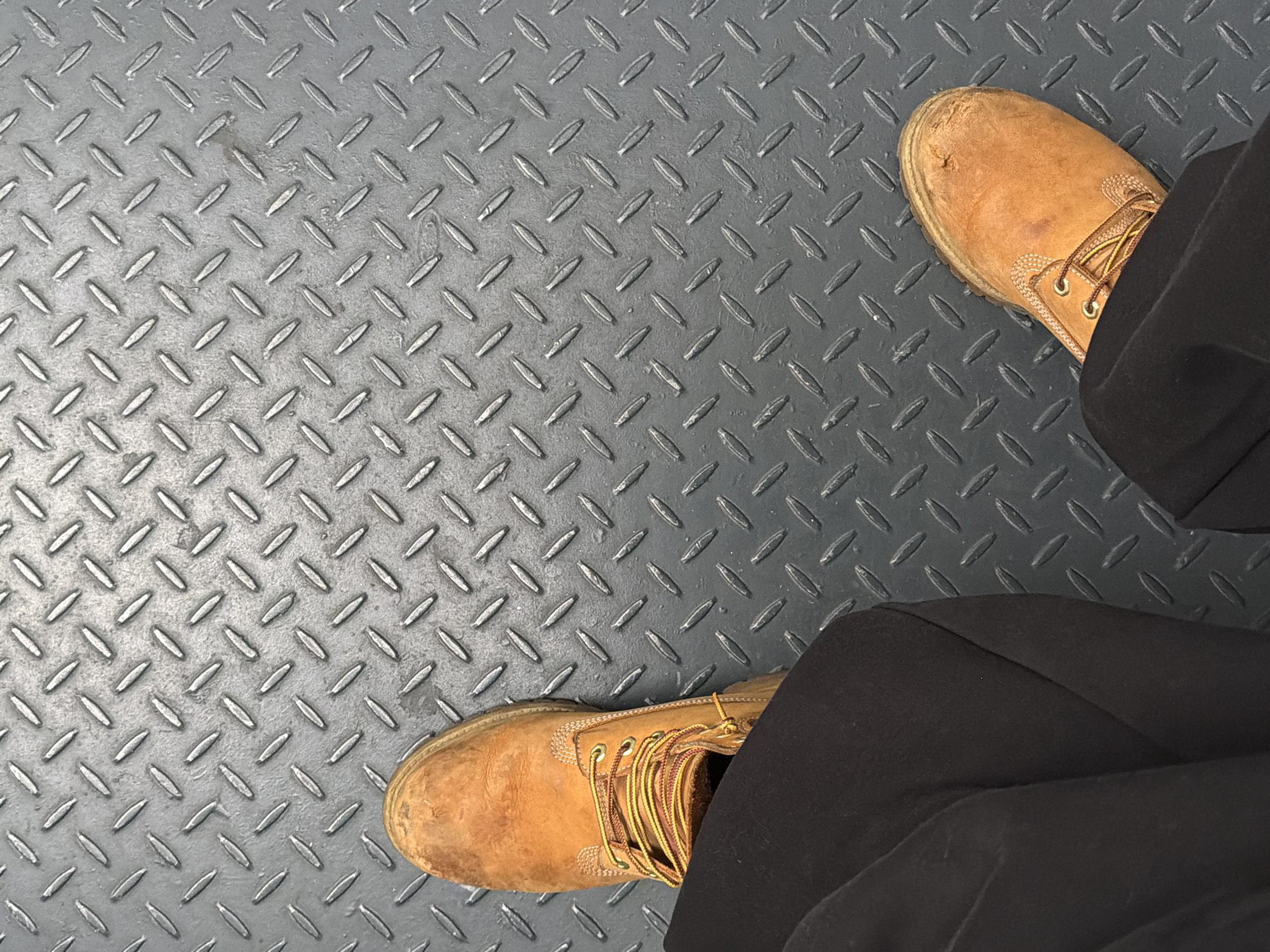 The image shows a person standing on a metal floor with a textured, repeating pattern of raised, elongated shapes. The person is wearing tan-colored work boots and black pants. The background is out of focus, but appears to be a building interior, given the flooring. There is no information in the image to indicate it's in London.