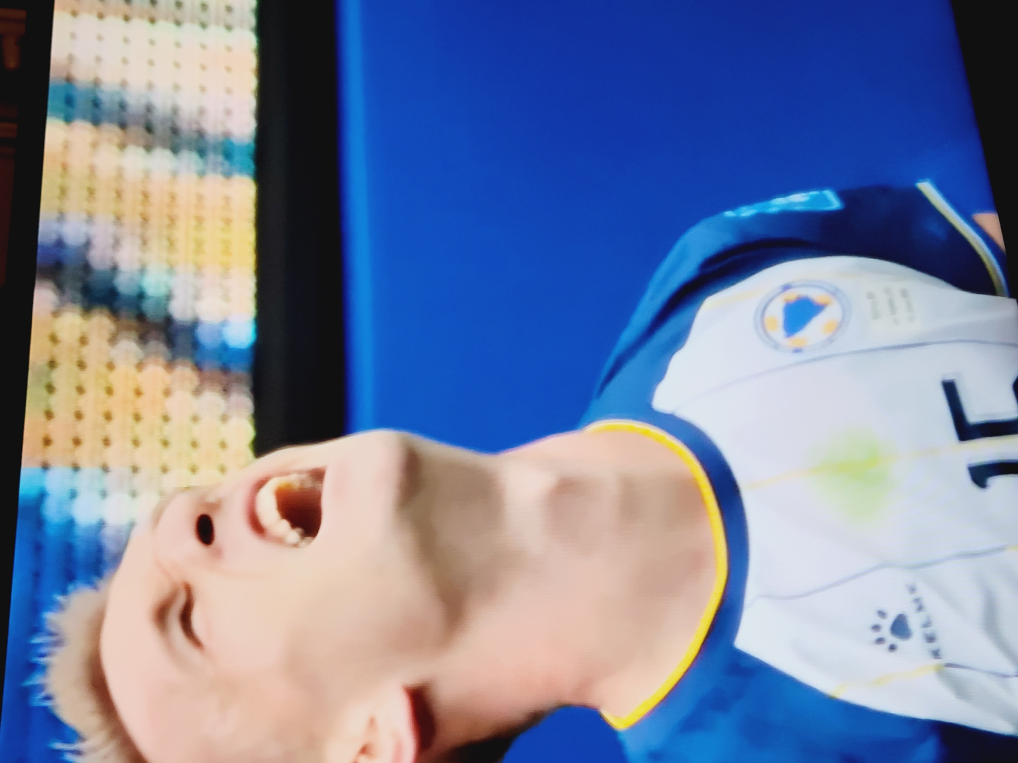The image shows a close-up of a male soccer player in a blue and white jersey with yellow trim. He is in mid-action, with his head thrown back and his mouth open in a scream or shout. The jersey has a logo on the left chest area, which appears to be a shield with a blue shape in the center, possibly a star or a country's emblem, surrounded by a yellow border. The word "KELME" and a paw print logo are visible on the jersey. The player is also wearing a yellow-trimmed collar. The background consists of a blurred display or scoreboard with rows of lights or pixels in yellow, blue, and white, and a solid blue surface below. The player's expression suggests intense emotion, possibly related to a game or event. No specific location in Baku City, Azerbaijan can be identified from the image alone.