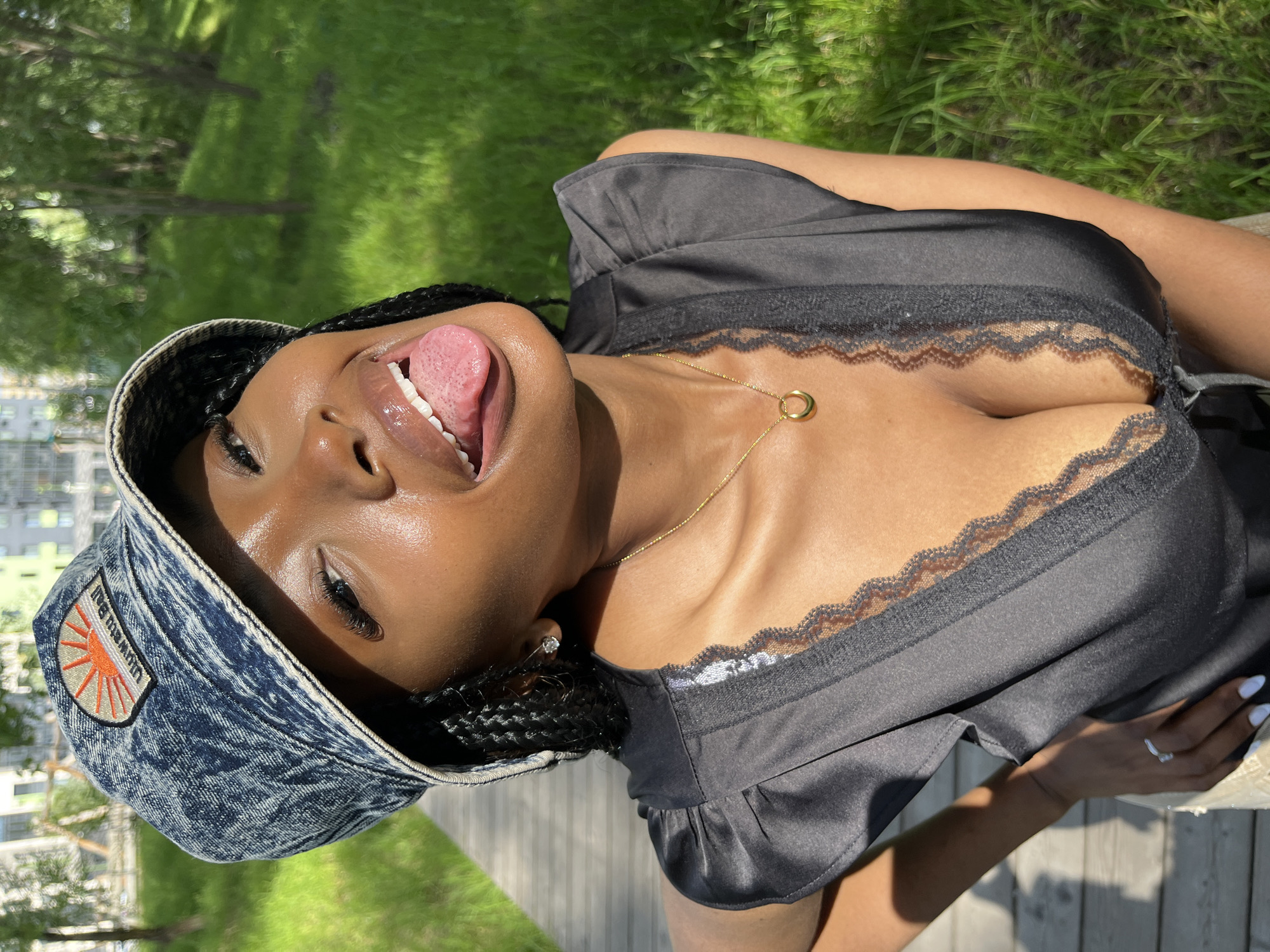 A young woman with dark skin, wearing a denim bucket hat and a black low-cut top with lace trim, is pictured outdoors. She is sticking her tongue out and smiling. She wears a gold necklace and a diamond stud earring. The background shows a grassy area and some buildings in the distance, suggesting an urban park setting. The image does not contain any information that would allow it to be located in the city of Kazan, Russia.