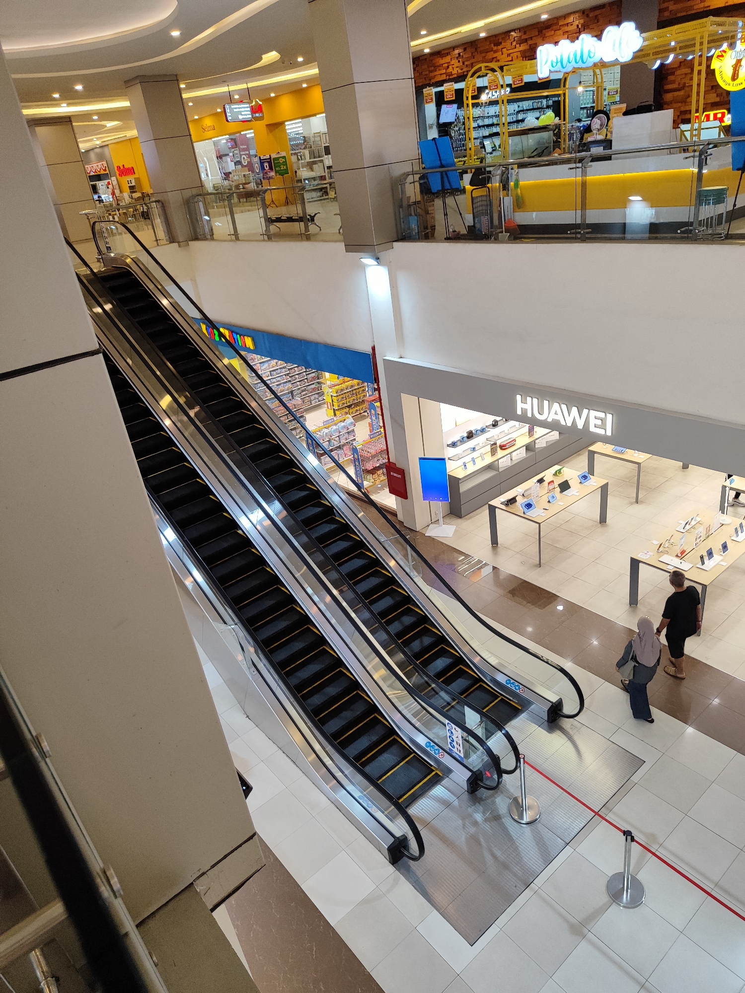 The image captures an interior view of a mall, likely in Sempusari, Indonesia, focusing on a section with escalators and storefronts. The scene is brightly lit by overhead lighting.

In the foreground, a set of parallel escalators descends, with black treads and silver railings. Two people are visible near the bottom of the escalators: a woman wearing a hijab and a long blue dress, and a man in a black t-shirt and shorts. They are walking away from the escalators. Red stanchions with a red rope cordon off an area in front of the escalators, creating a pathway.

Further back, a store with the "HUAWEI" logo is prominent. This store displays various electronic devices on tables, including what appear to be smartphones and laptops. Across from the Huawei store, another shop with a yellow facade and the sign "Potatoo Lab" is visible. This area appears to be a kiosk or smaller retail space, with merchandise displayed behind a counter. Above and to the left, a store with a yellow facade and the sign "Selma" can be seen, also with merchandise displayed within.

The mall's architecture includes white pillars and a white ceiling with recessed lighting. The floor is tiled with a light-colored, patterned tile. The overall impression is a modern, commercial indoor environment.