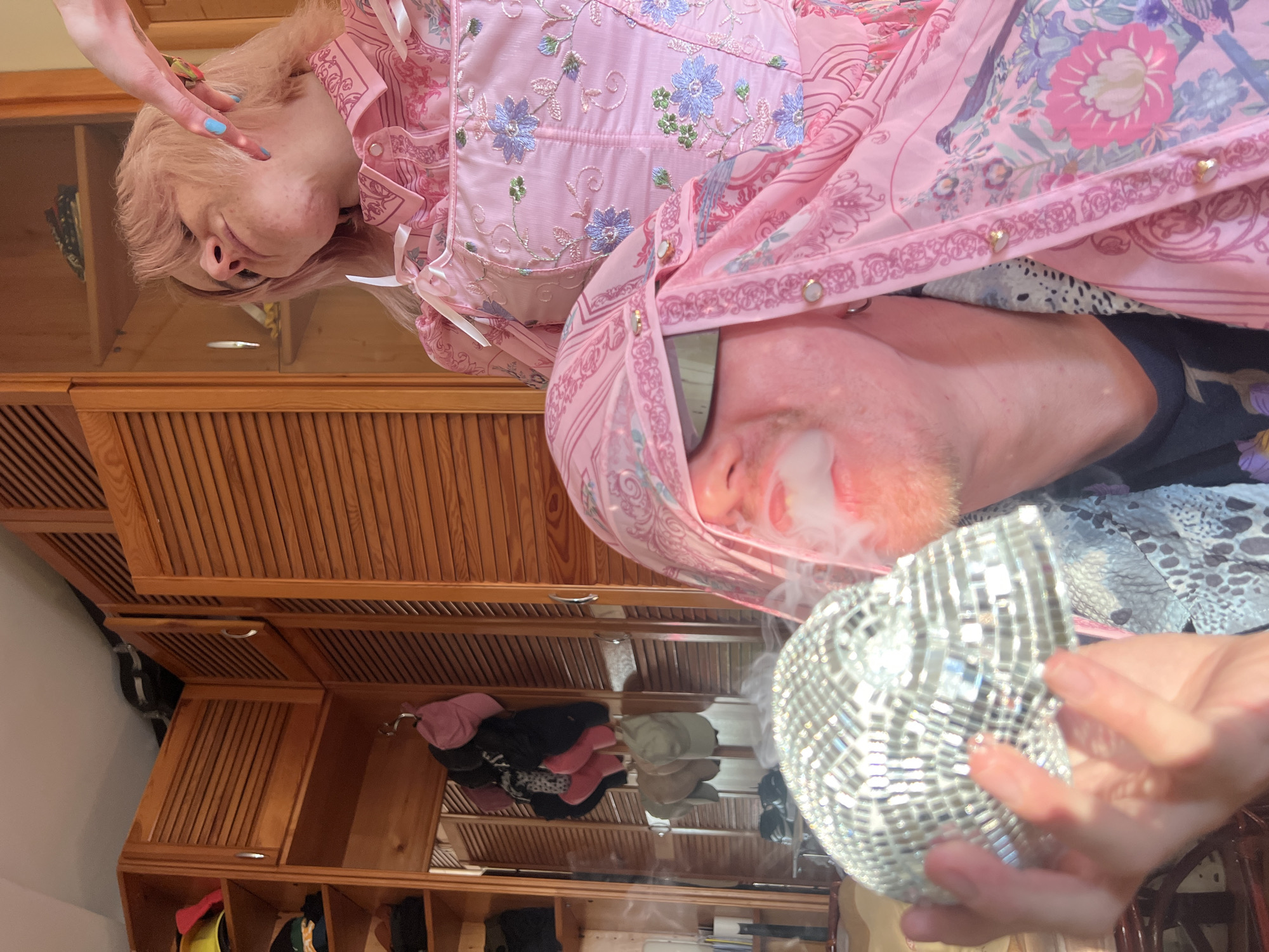 The image shows a mirrored reflection of two people inside a room, likely a home interior. One person with pink hair and pale skin is wearing a pink outfit with floral patterns and white ties. Their fingers with blue nail polish are visible near their face. The second person, with a reddish-orange beard and pale skin, is also wearing pink clothing. They are holding a disco ball and exhaling smoke, possibly from a vaping device or cigarette. The reflection is on a wooden surface, likely a table or dresser top. The background reveals wooden cabinets or storage units, with various items stored inside. No specific details suggest a location in Belgrade, Serbia.