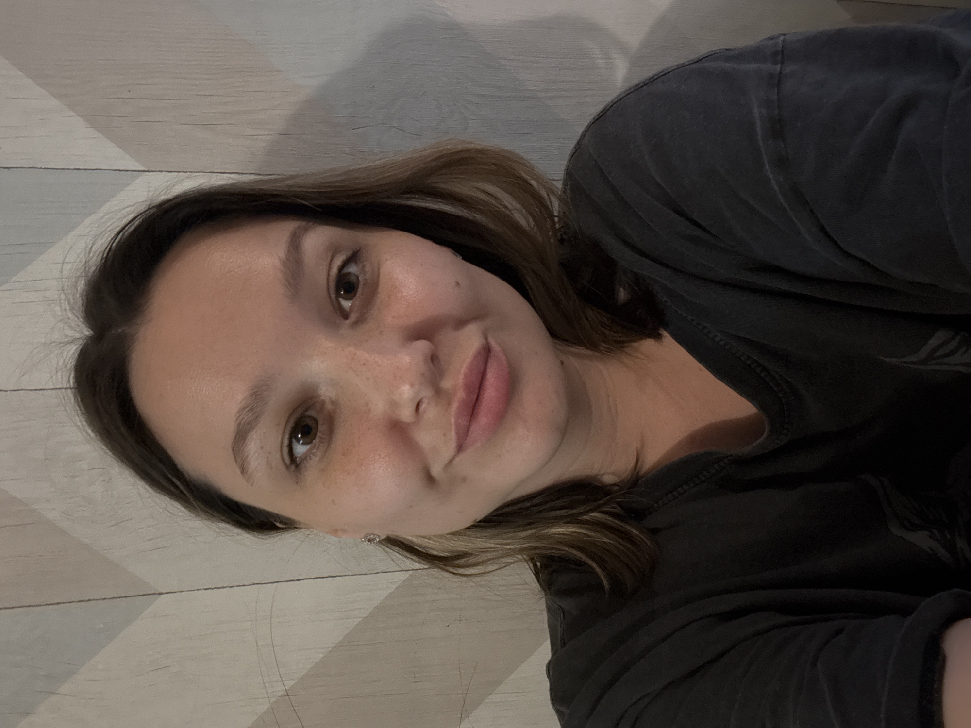 A medium shot shows a woman with brown hair and brown eyes smiling at the camera. She is wearing a dark gray t-shirt. The background is a wall with a diagonal pattern of light and dark gray. There is no information about the location being in the city of Белоус, Russia.