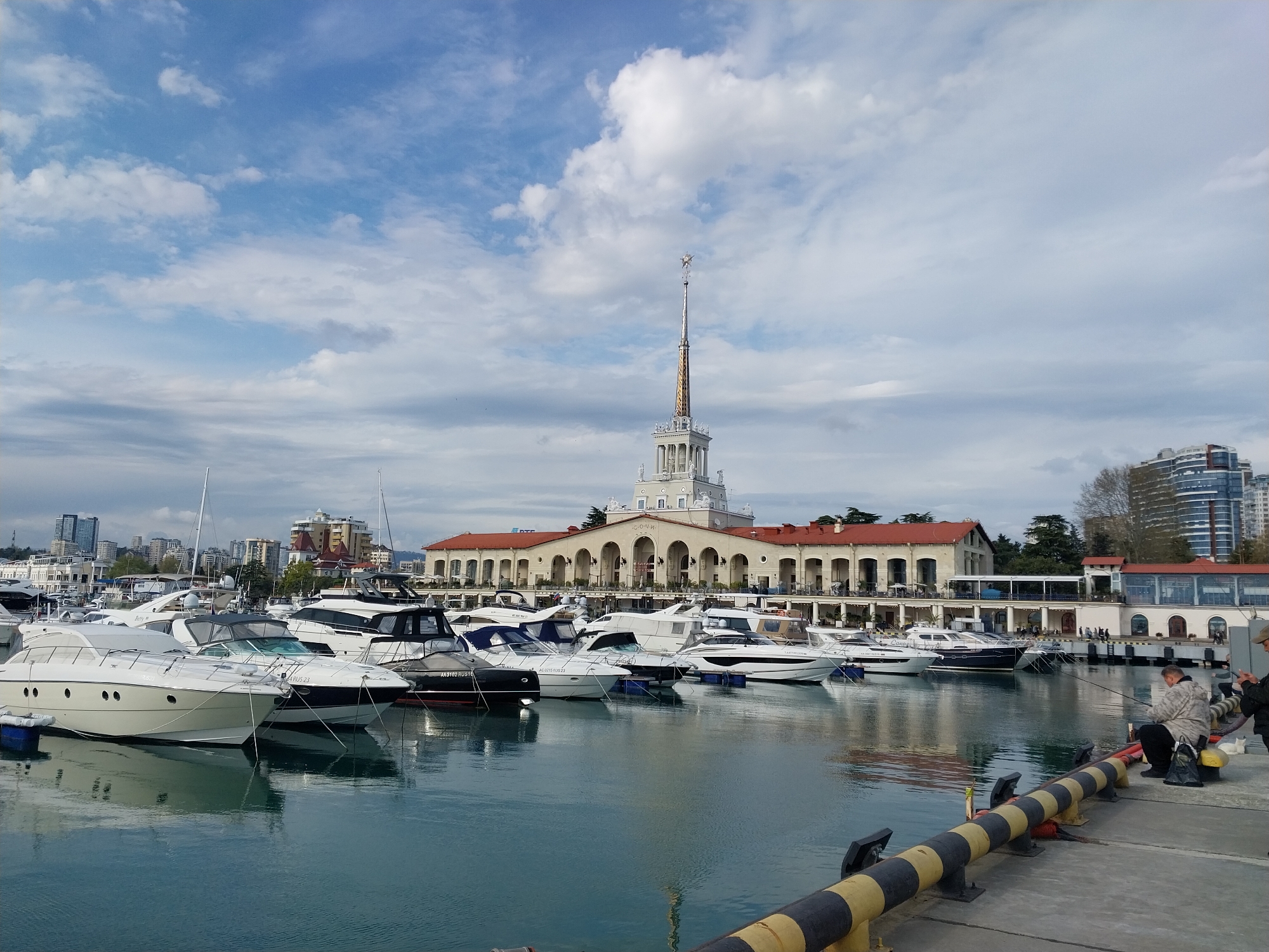 This image captures a picturesque marina scene in Sochi, Russia, under a partly cloudy sky. The foreground features calm, turquoise water reflecting the sky and docked vessels, with a pier section in the lower right displaying distinctive black and yellow striped bollards.
Numerous yachts and motorboats of various sizes, predominantly white, are moored along the docks, their sleek hulls and cabins creating a dynamic pattern on the water. Registration numbers such as "AK 3102 RUS 16" and "AK 0215 RUS 23" are visible on the sterns of some boats.
The central and most prominent structure is a grand, classical-style building, likely the Sochi Sea Terminal, characterized by multiple arches, columns, and a red-tiled roof. A very tall, slender spire, topped with a star-like ornament, rises majestically from its center. The word "СОЧИ" (Sochi) is faintly discernible above one of the building's main archways. In the background, modern city buildings rise, indicating an urban setting surrounding the port.
On the right side of the pier, two men are visible. One is seated, appearing to be engaged in fishing or simply relaxing, while the other stands beside him. The scene suggests a peaceful daytime environment, with bright natural light diffused by scattered white clouds. The overall atmosphere is one of calm leisure within a bustling port city.