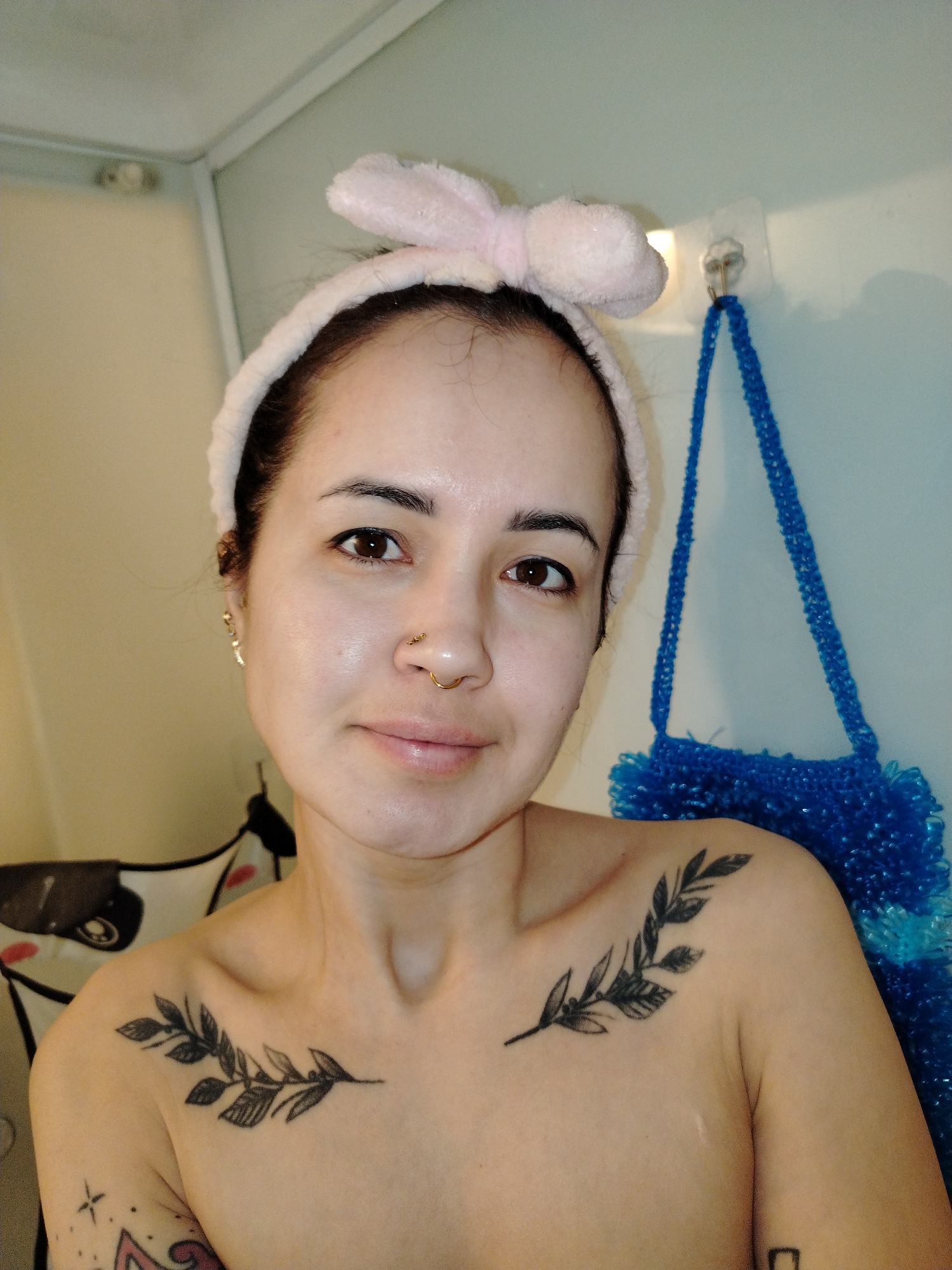 A woman with fair skin and dark hair pulled back by a light pink headband with bunny ears poses for a close-up shot. She has brown eyes, a nose ring, and several tattoos visible on her chest and arm. Behind her, a blue loofah hangs from a hook on a light-colored wall. The setting appears to be a bathroom. The location is not discernible from the image and cannot be identified as Noviy, Russia.