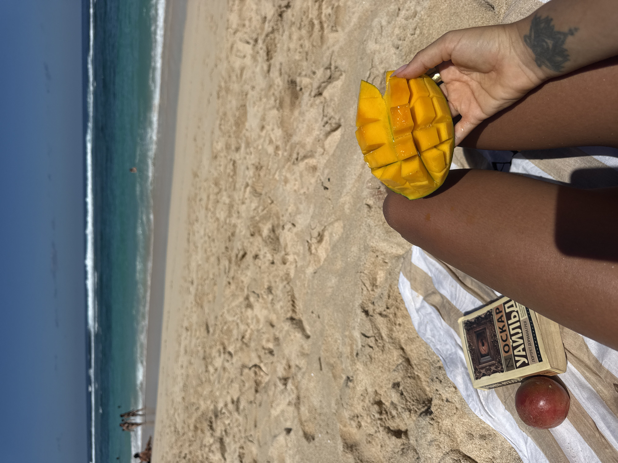 A tanned person sits on a striped towel on a sandy beach in Kuta Selatan, Indonesia, holding a peeled and cubed mango in their hand. A book titled "Oscar Wilde" in Russian rests near their legs along with a passion fruit. The ocean and a blue sky are visible in the background, and people are visible along the shoreline. The person has a tattoo on their left wrist.