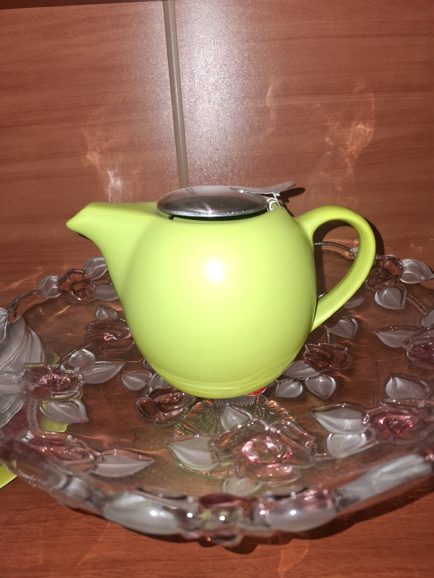 The image shows a bright yellow teapot with a silver lid resting on a clear glass decorative plate. The plate has a floral design with pink and clear glass elements. The teapot and plate are placed on a reddish-brown surface that appears to be a countertop or table with a wood grain pattern. The background is a textured reddish-brown wall, possibly made of tile or laminate, with a vertical seam running down the center. There are reflections and light glares on the surfaces of the objects and the background, suggesting indoor lighting. No people are visible in the image. The location is not specified in the image, and therefore, it cannot be determined to be in Almaty, Kazakhstan.