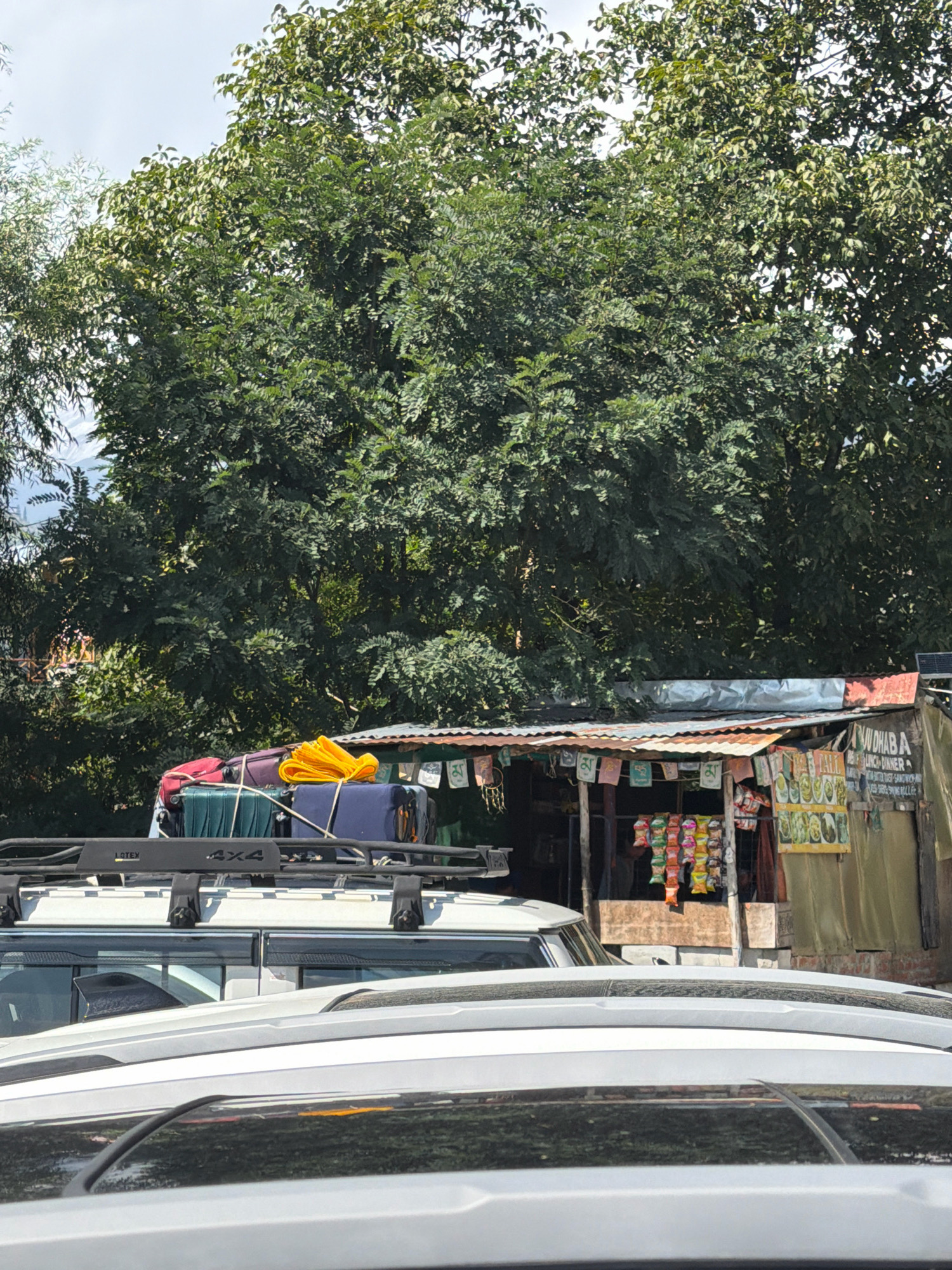 In Vashisht, India, a roadside dhaba, likely a food stall, is visible under a corrugated metal roof. The dhaba displays various snacks and colorful food items, suggesting it is open for business. Above the dhaba, a rack on a white SUV is laden with luggage, including suitcases and a bright yellow tarp. The scene is framed by lush green trees and a partly cloudy sky. The foreground shows the rooflines of other vehicles, indicating a traffic situation or parking area. The overall impression is a snapshot of roadside commerce and travel.