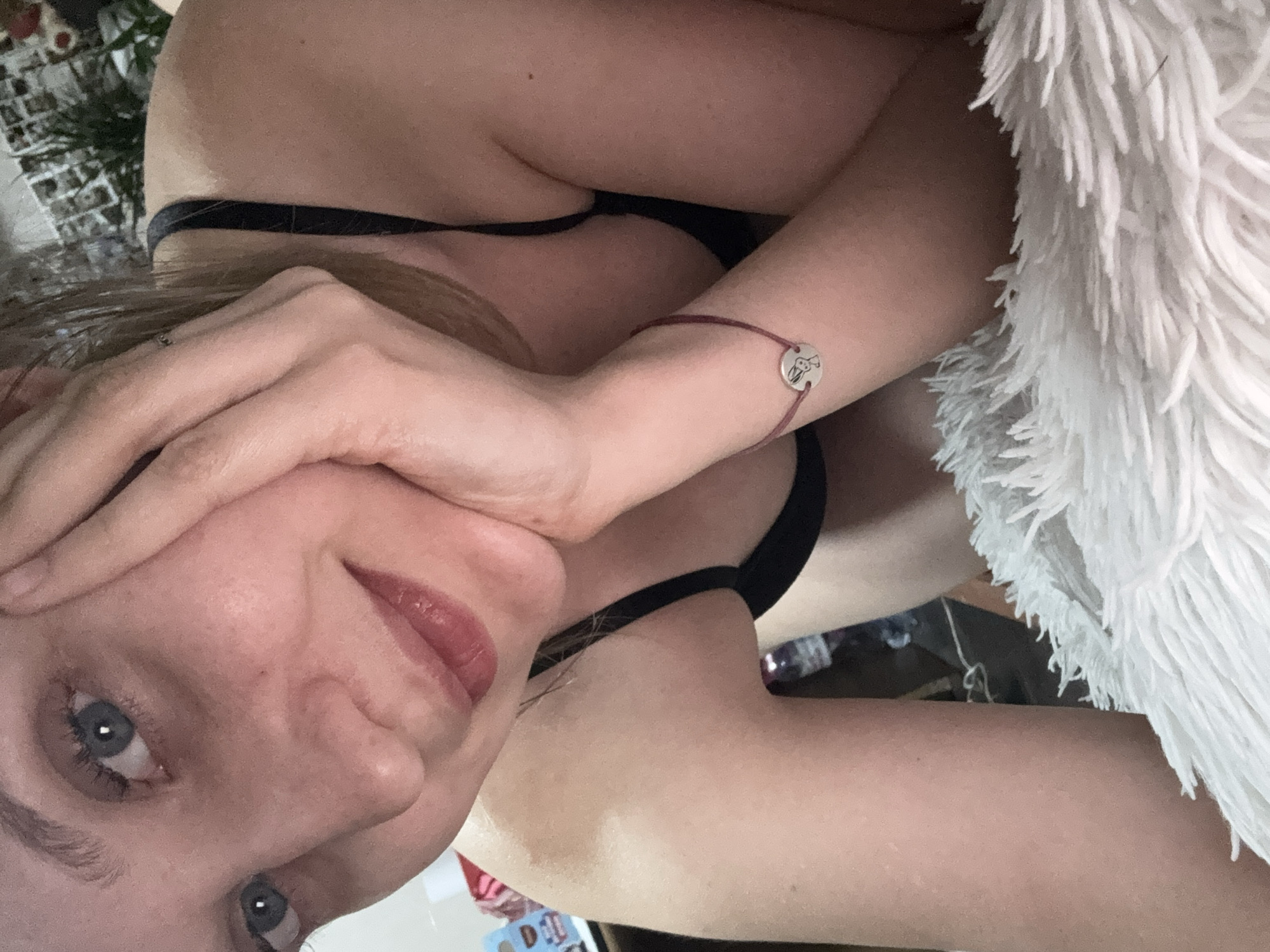 The image is a close-up, indoor selfie of a young woman. She is lying down, resting her head on her hand, and looking towards the camera with a slight smile. She is wearing a black bra. A thin, burgundy bracelet with a circular charm depicting a rabbit is visible on her wrist. The background is blurred and suggests an indoor setting with some objects on shelves and a plant. There is no information in the image that would indicate its location in Izhevsk, Russia, or any other specific city.