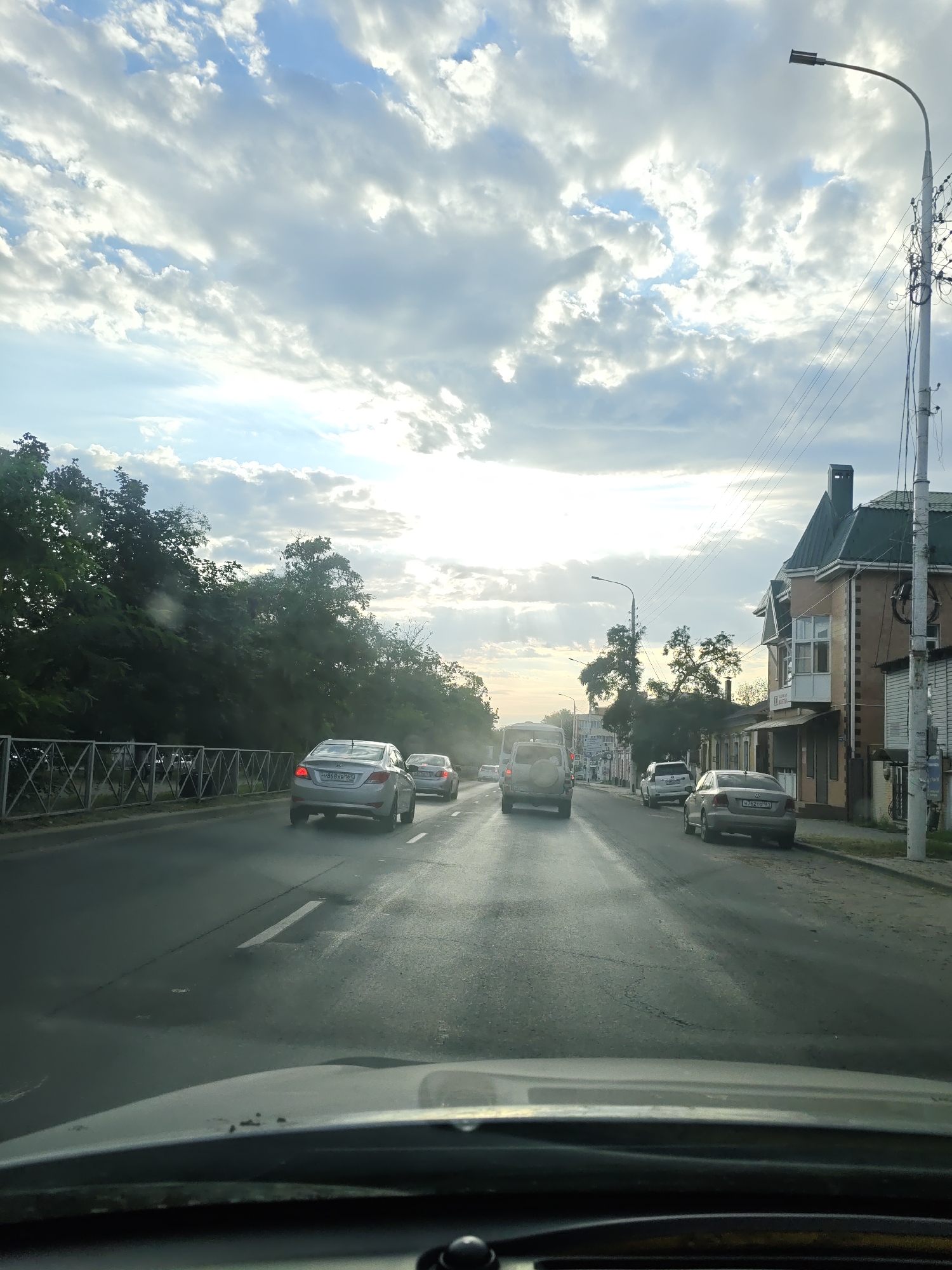The image depicts a street scene in Novocherkassk, Russia, on a cloudy day with shafts of sunlight breaking through. The perspective is from inside a vehicle, looking forward. Several cars are traveling on a paved road. On the left side of the road, there are trees and a metal fence. On the right side, there are buildings, including a two-story brick building with a green roof, and a street lamp. The sky is filled with dramatic clouds.
