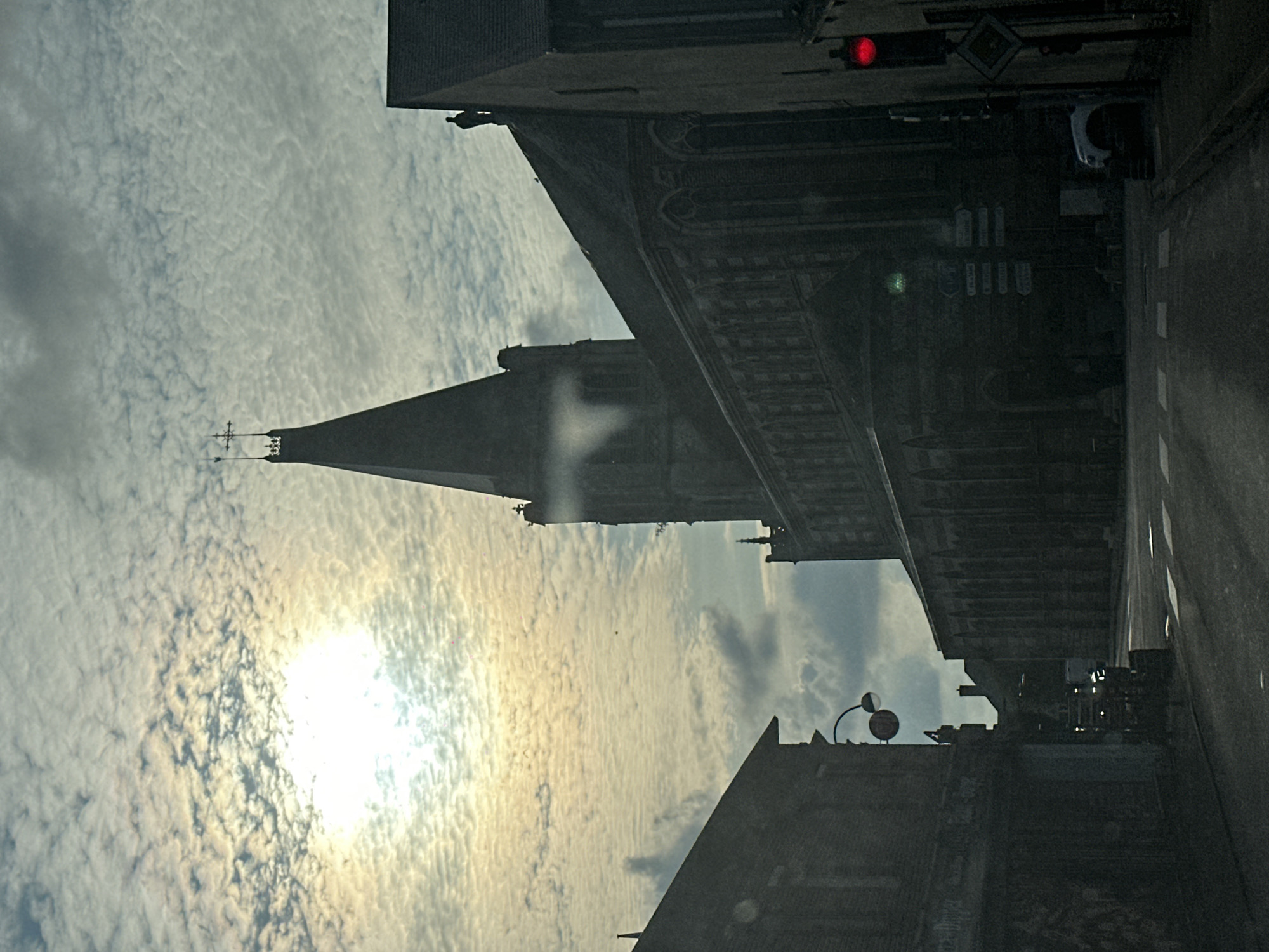 The image depicts a street scene in Saint-Maclou-de-Folleville, France, dominated by the silhouette of a large church with a tall spire. The sun is low in the sky, partially obscured by textured clouds, casting a diffused light that creates a somewhat muted and dramatic atmosphere. In the foreground, the dark facades of buildings line the street, one of which appears to be a shop with faded lettering. The road in front of the church has crosswalk markings and appears wet or reflective. A red traffic light is visible on the right side of the frame. There is a white car parked on the right side of the road, and a vehicle that could be a truck or van is visible further down the street in the center. A person is visible on the roof of a building to the right of the church spire, and another figure stands near the top of the church's spire. Signposts are affixed to the wall of the church.