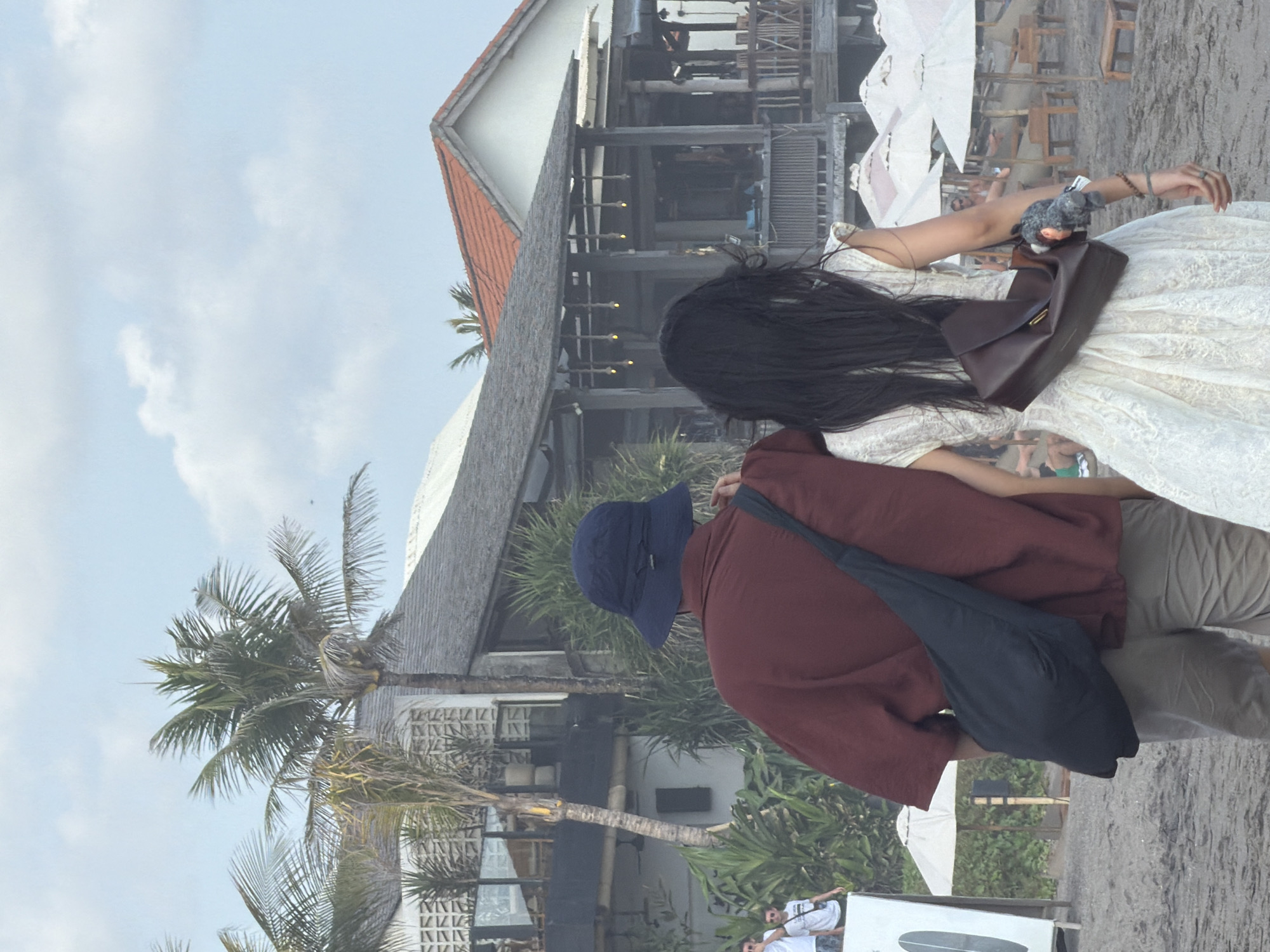 The image shows a couple walking on a beach in North Kuta, Indonesia. The man is wearing a maroon shirt, khaki shorts, and a blue bucket hat. He has a black bag slung across his body. The woman is wearing a white lace dress and has a brown handbag slung across her body. They are walking away from the camera. In the background, there are buildings with thatched roofs and palm trees. There are also several white beach umbrellas and lounge chairs. The sky is overcast with some clouds.