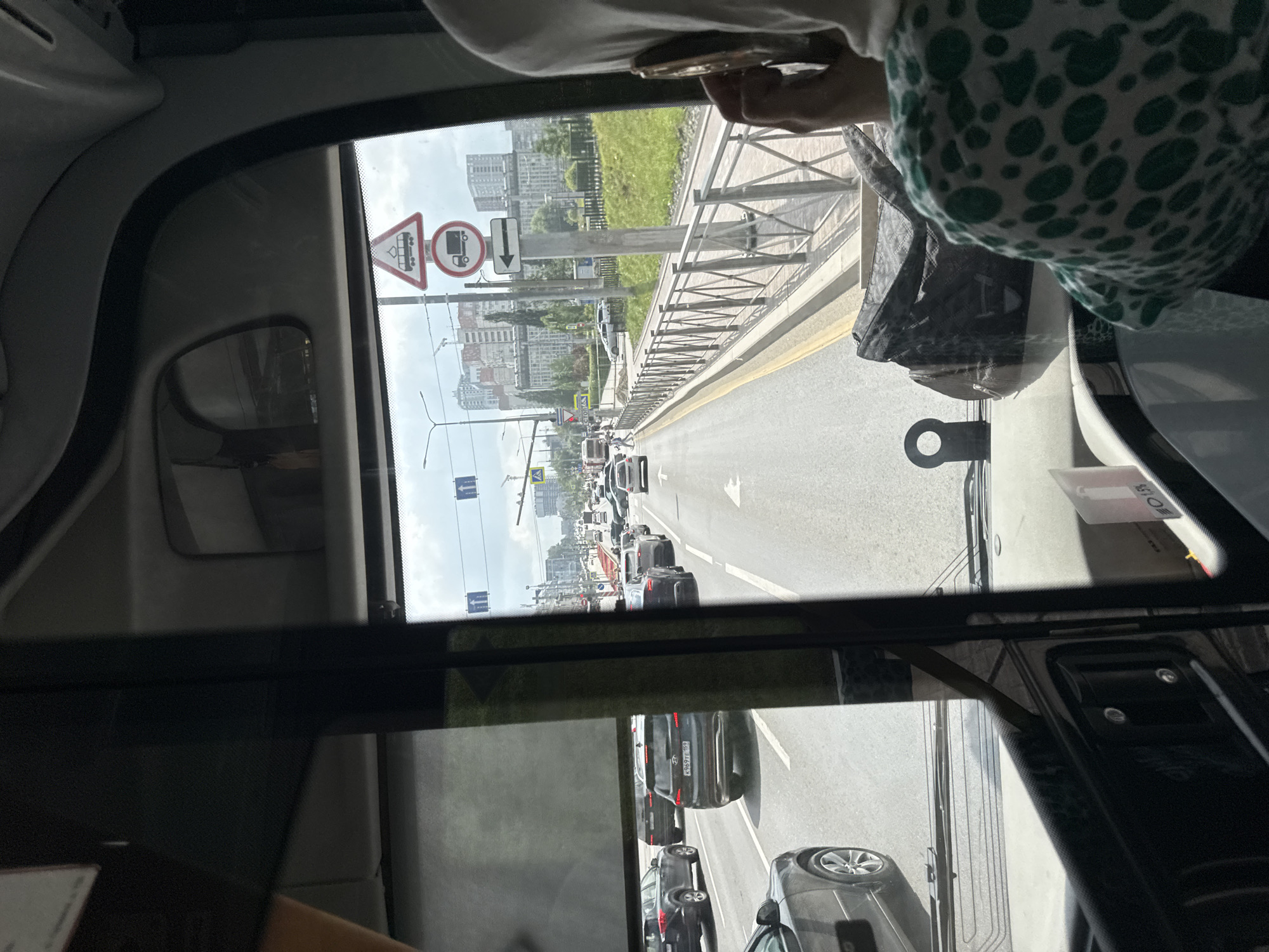 The image is taken from the interior of a bus looking forward through the windshield. The bus is in traffic on a multi-lane road in a city. There are numerous cars in front of the bus, suggesting a traffic jam. The road has lane markings and sidewalks visible. On the right side of the road, there is a metal fence and a grassy embankment with trees and buildings in the background. In the distance, there are tall apartment buildings and construction cranes, indicating an urban environment.
Several traffic signs are visible. One sign is a triangular sign with a trolleybus symbol inside a red border, indicating a trolleybus route. Below that is a circular sign with a truck symbol inside a red border, indicating a prohibition for trucks. There is also a rectangular sign with a white arrow pointing left, indicating a direction. These signs are consistent with traffic regulations and road infrastructure found in cities.
On the right side of the bus, a person wearing a white hijab and a green and white patterned shirt is visible. This person is holding a mobile phone to their ear, possibly making a call.
The overall scene depicts a typical urban traffic situation on a bright, cloudy day. Given the presence of trolleybus signs and the architectural style of the buildings, this scene is consistent with a location in Perm, Russia. Specifically, the presence of trolleybus infrastructure is a characteristic feature of Perm's public transportation system.