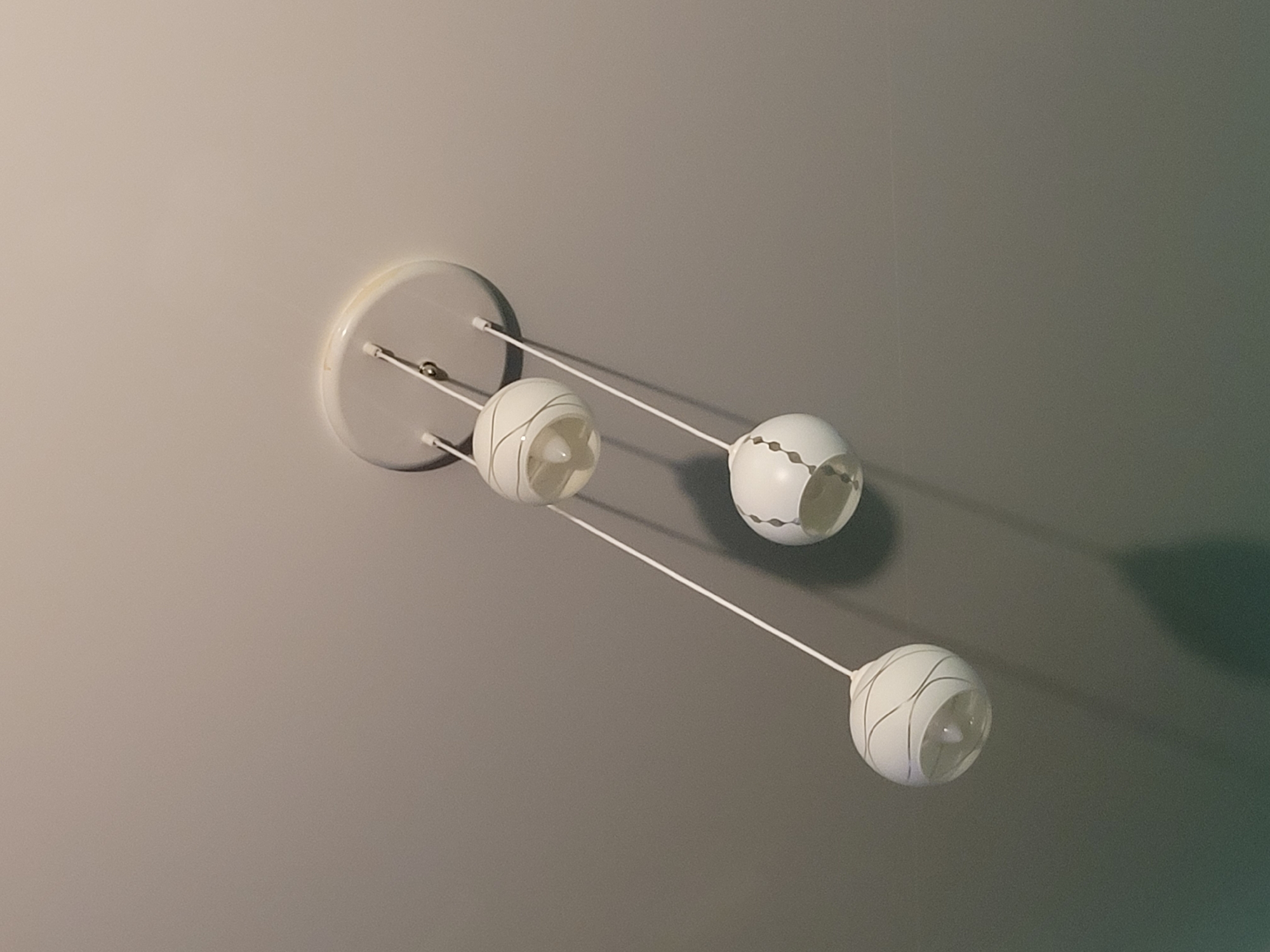 The image shows a white, three-bulb ceiling light fixture against a gray ceiling. The fixture consists of a circular base mounted to the ceiling, from which three arms extend, each terminating in a globe-shaped lamp. The lamps are also white with curved decorative lines. The scene appears to be indoors. The image does not depict any people, actions, or interactions. The location in the city of Izhevsk, Russia is not identifiable from the image.