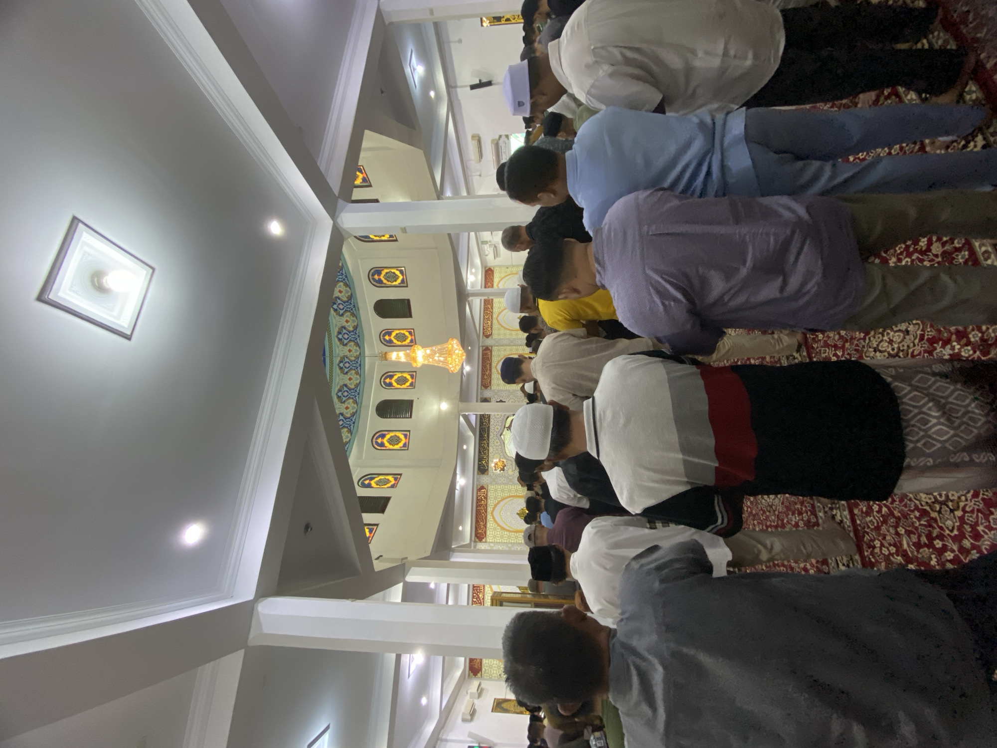 The image shows a gathering of Muslim men in a mosque, likely for prayer, in Lhokseumawe, Indonesia. The men are facing forward, and many are wearing white skullcaps. The mosque interior features high ceilings with ornate decorations, including a chandelier and stained-glass windows. The floor is covered with a red patterned carpet. The overall atmosphere appears to be one of reverence and community.