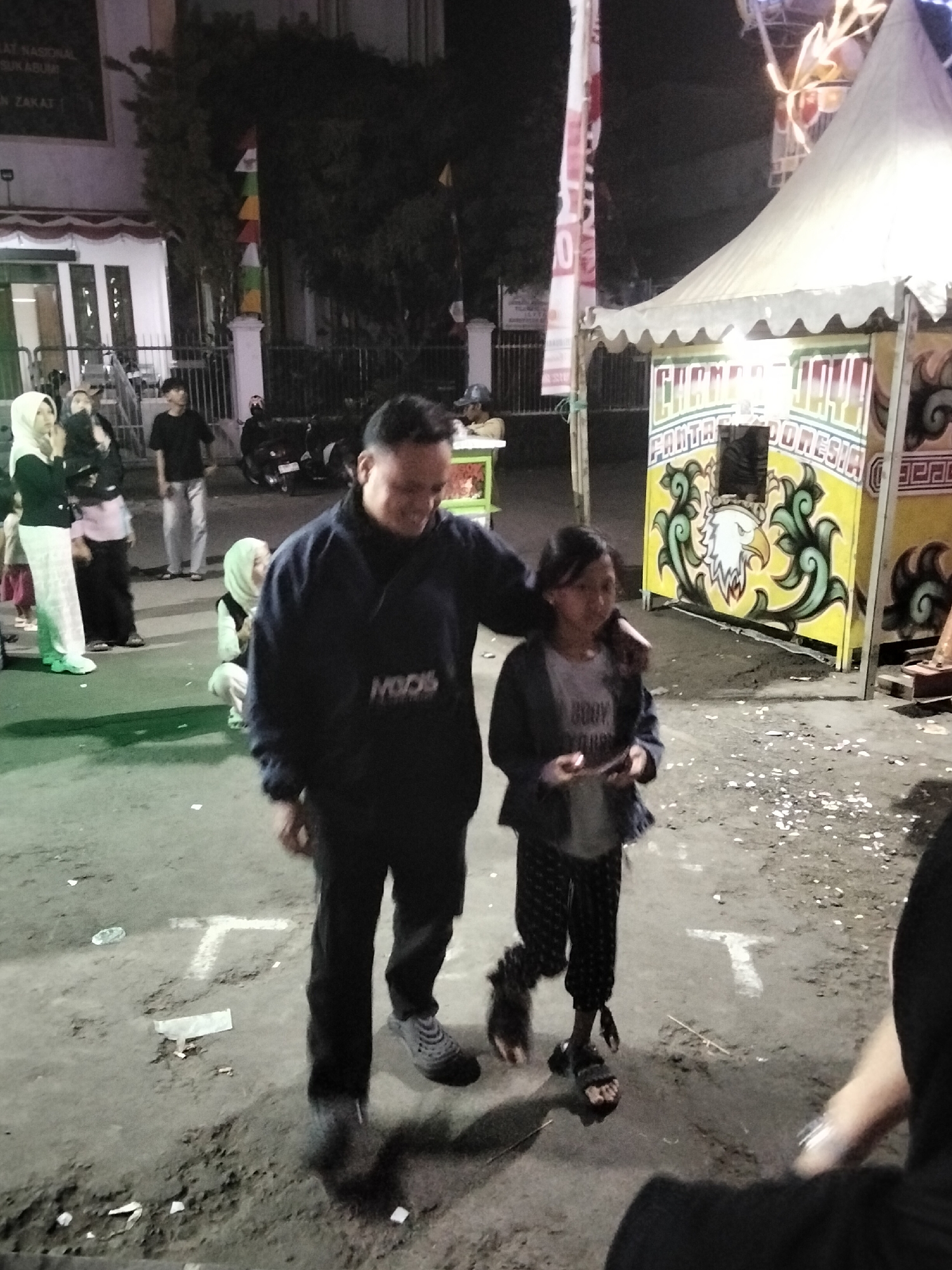 In Cisaat, Indonesia, a nighttime street scene unfolds near a tented stall adorned with yellow and green imagery of an eagle and decorative elements. A man in a dark blue jacket and black pants walks with a young girl in a grey shirt and patterned pants. The girl is holding a small object. Other individuals, including a woman in a white hijab and a man in a black t-shirt, are visible in the background near a building. A motorcycle is parked on the left side of the image. The ground appears to be unpaved and dusty, with scattered white debris. A banner hangs vertically on the left side of the frame.