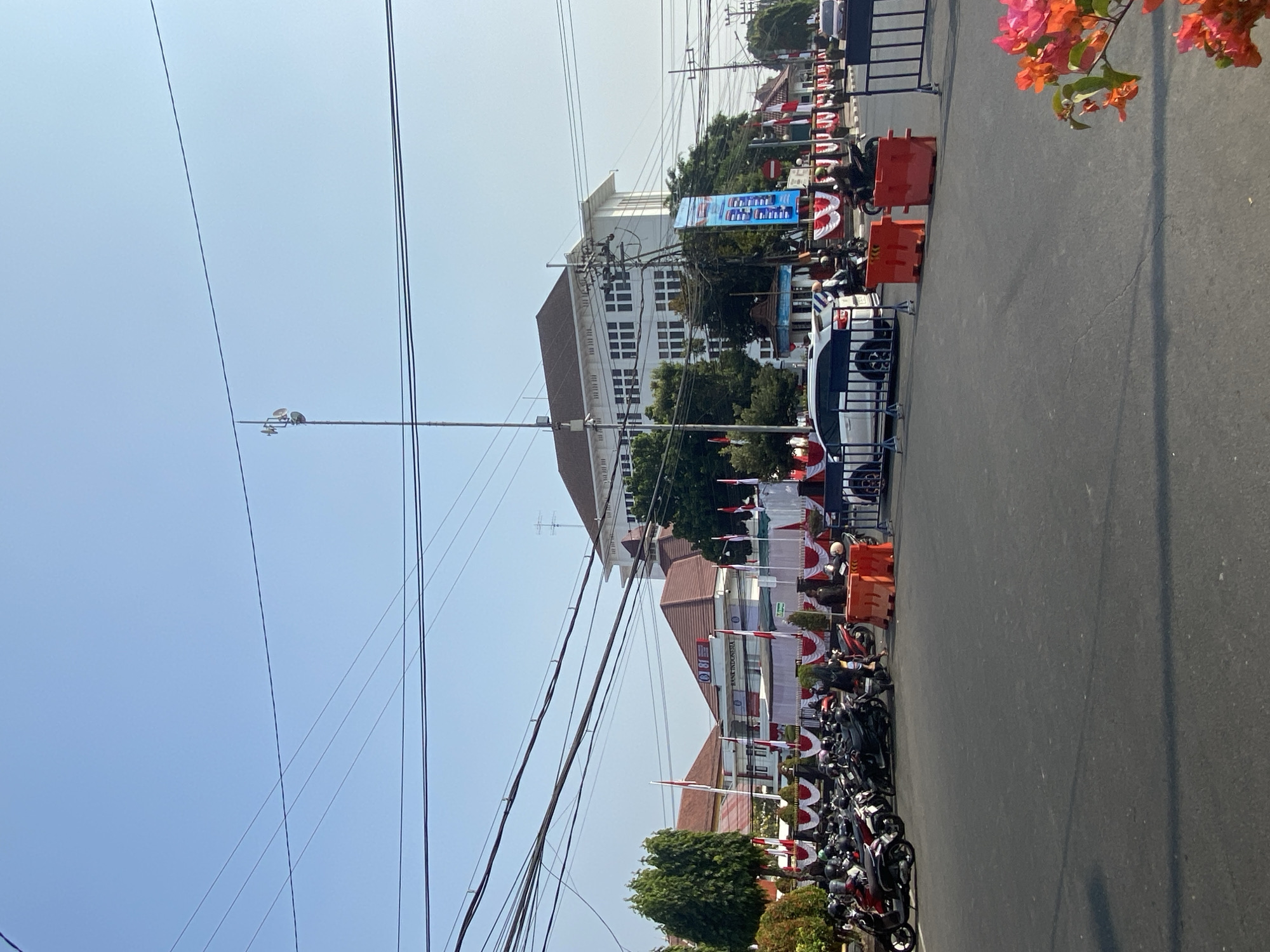 A clear, daytime shot captures a street scene in Kediri City, Indonesia. In the foreground, a paved road shows some parked motorcycles and a few vehicles. In the middle ground, a row of parked motorcycles is visible on the left, with a white car and several motorcycles in motion on the right. Beyond the road, a building adorned with flags and banners is situated. This building appears to be "Bank Indonesia." The background features a large, white building with multiple windows and a rooftop. Overhead, a network of power lines crisscrosses the sky. The overall impression is a bustling urban environment under a bright sky.