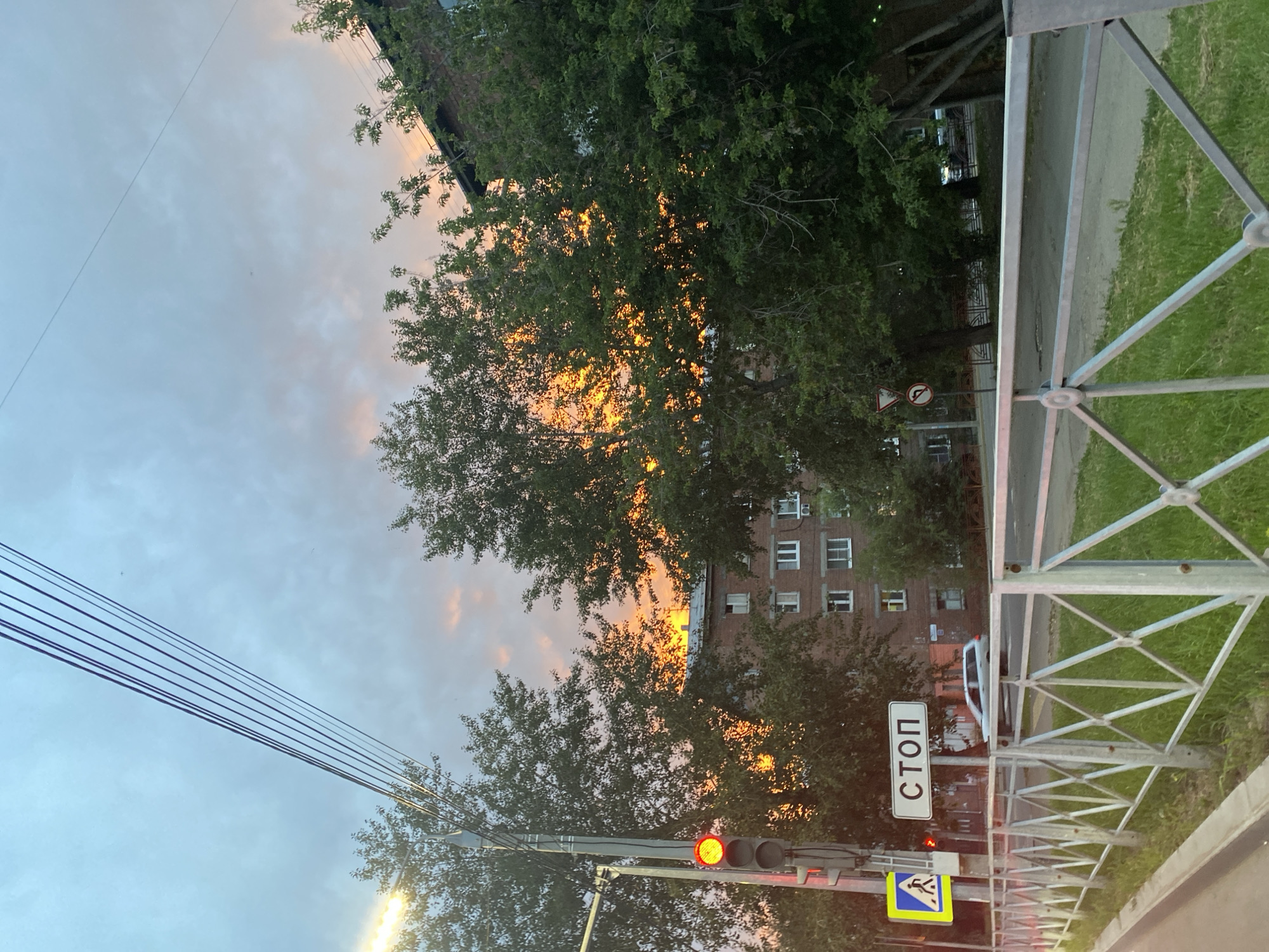 The image captures a street scene in Irkutsk, Russia, during sunset. The sky is a mix of blue and grey clouds with vibrant orange and yellow hues indicating the setting sun, which is partially obscured by trees. A traffic light with a red illuminated signal is visible on the left, next to a pedestrian crossing sign. A "СТОП" (STOP) sign is prominently displayed on a pole. Trees line the street, with a brick apartment building visible in the background. A silver car is driving on the road, and a metal fence with a diamond pattern runs along the sidewalk. The overall atmosphere is that of an urban environment at dusk.