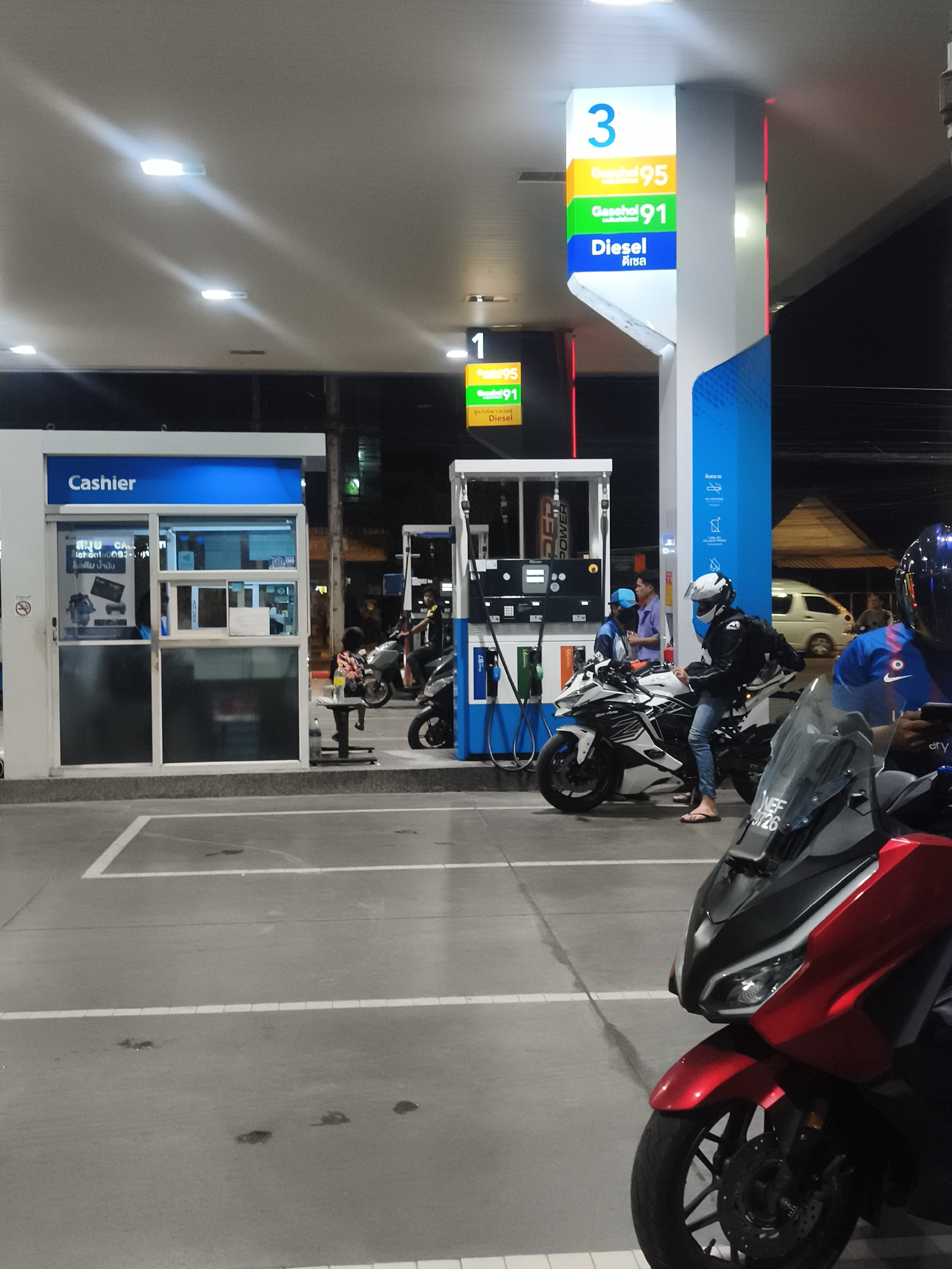 The image captures a bustling gas station at night in Baan Mae Nam, Thailand, under bright artificial lights. The primary activity appears to be fueling motorcycles.
In the foreground on the right, a red scooter, possibly a Honda Forza, with "WEF 9726" visible on its windshield, is parked, partially obscuring a person in a blue shirt and dark helmet. Central to the frame, a person wearing a white helmet, black jacket, and jeans stands next to a white sportbike (Kawasaki Ninja ZX-25R), seemingly preparing to refuel. Behind them, a gas station attendant in a light blue shirt and cap is visible near the pump. Further back, another person on a scooter and a white van are partially visible.
To the left, a "Cashier" booth is brightly lit, featuring an open service window. A small child sits on a stool near the cashier booth. Various signs indicate fuel types and pump numbers. A prominent vertical sign displays "3" at the top, followed by "Gasohol 95," "Gasohol 91," and "Diesel" (with "ดีเซล" in Thai below it). Another smaller sign to the left shows "1," "95," "91," and "ซูเปอร์พาวเวอร์ Diesel." The pumps themselves bear the brand name "POWER." Text on the cashier booth window includes "สมุย CAL" and "ไม่เดิม นํ้ามัน," possibly a specific instruction or warning. The ground is concrete with white parking lines, some marked by small dark stains. The scene is well-lit by overhead rectangular lights.