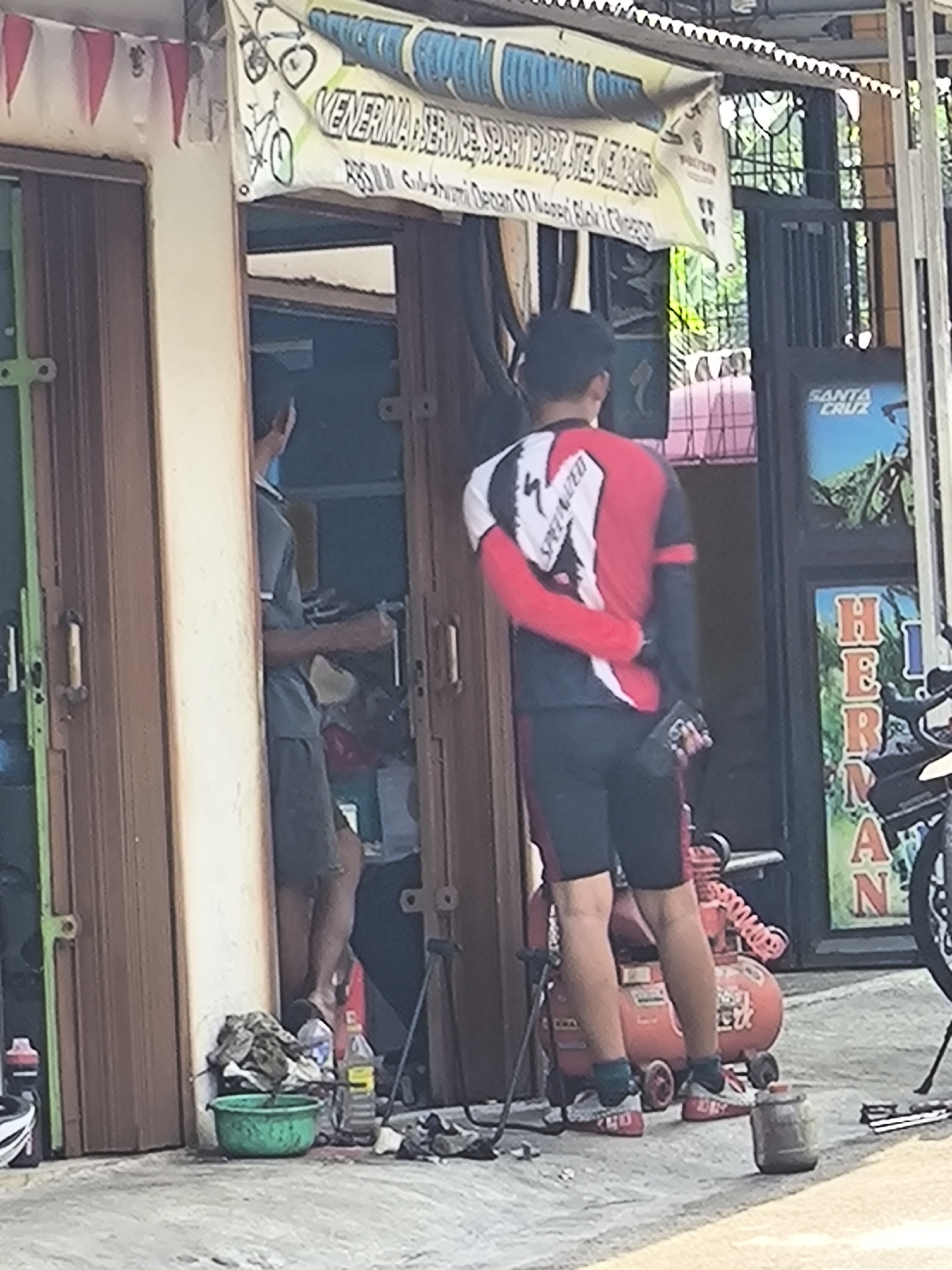 The image captures a street-level view of a bicycle repair shop in Cilegon, Indonesia. In the foreground, a person wearing a red and black cycling jersey and black shorts stands with their back to the camera, holding a smartphone in their right hand. They are wearing red cycling shoes with white laces and blue socks. To the left of this individual, an open doorway reveals another person in a dark grey uniform, possibly an employee, standing inside the shop and holding a light-colored object.
In front of the shop entrance, a green basin filled with water and some cloth is visible on the ground. Next to it, a large red air compressor with coils and wheels sits on the pavement. Behind the air compressor, part of a motorcycle is visible.
Above the doorway, a banner is displayed, featuring text that appears to be in Indonesian. The banner depicts a drawing of a bicycle and the words "MENERIMA SERVICE... SPARE PART..." which suggests the shop offers repair services and sells spare parts for bicycles.
Further to the right, a black metal gate encloses the area. A poster with the "SANTA CRUZ" logo and a depiction of a cyclist is mounted on the wall. Below this, another sign with vertical text spelling out "HERMAN" is partially visible. The overall environment appears to be an urban setting, with pavement and building structures.