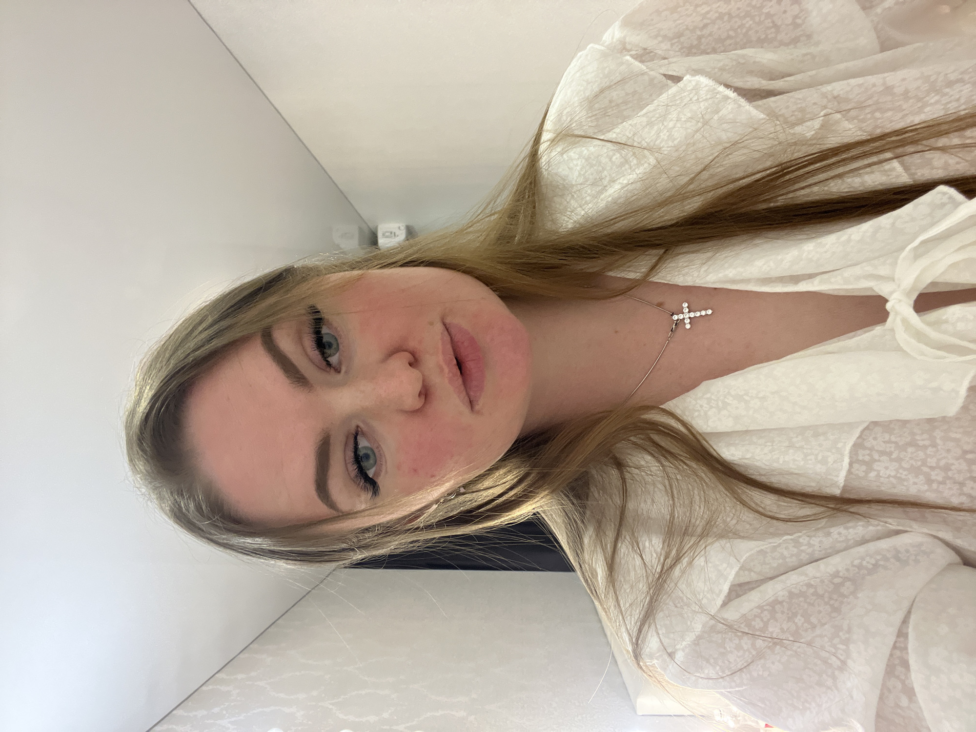 The image captures a close-up portrait of a young woman in an indoor setting in Sevastopol, Ukraine. She has long, light brown hair that cascades over her shoulders and chest, partially obscuring her white, ruffled blouse. Her light blue eyes are a central feature, accentuated by visible eyeliner and mascara, complemented by well-defined eyebrows. Her lips are full and slightly parted, contributing to a neutral or subtly contemplative expression as she looks directly at the camera.
She is wearing a white blouse made of a light, possibly sheer fabric with a delicate, small floral or dotted pattern. Around her neck, a silver chain necklace displays a sparkling, diamond-encrusted cross pendant. The background consists of plain, light-colored walls and possibly a ceiling line in the upper left corner. A light-colored surface, possibly a countertop or table, is visible in the foreground beneath her face. The lighting suggests a well-lit indoor environment. There is no visible text in the image.