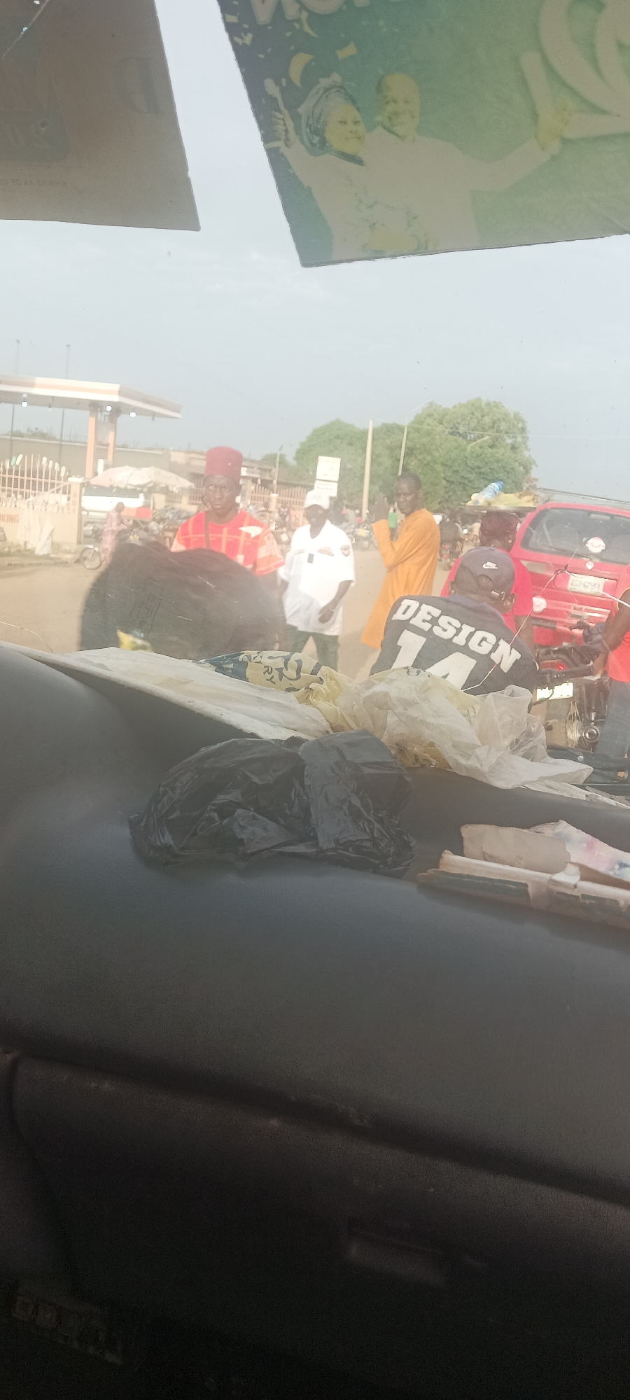 The image shows a street scene in Mangu Town, Nigeria, as viewed from inside a vehicle. In the foreground, the vehicle's dashboard is cluttered with items including plastic bags and rolled-up papers. Through the windshield, several people are visible on the street. One man wears a traditional red patterned outfit and hat. Another individual wears a white uniform. A third man is dressed in an orange robe. A red car is visible in the background, along with other vehicles, pedestrians, and a fuel station. The overall scene indicates a busy urban environment.