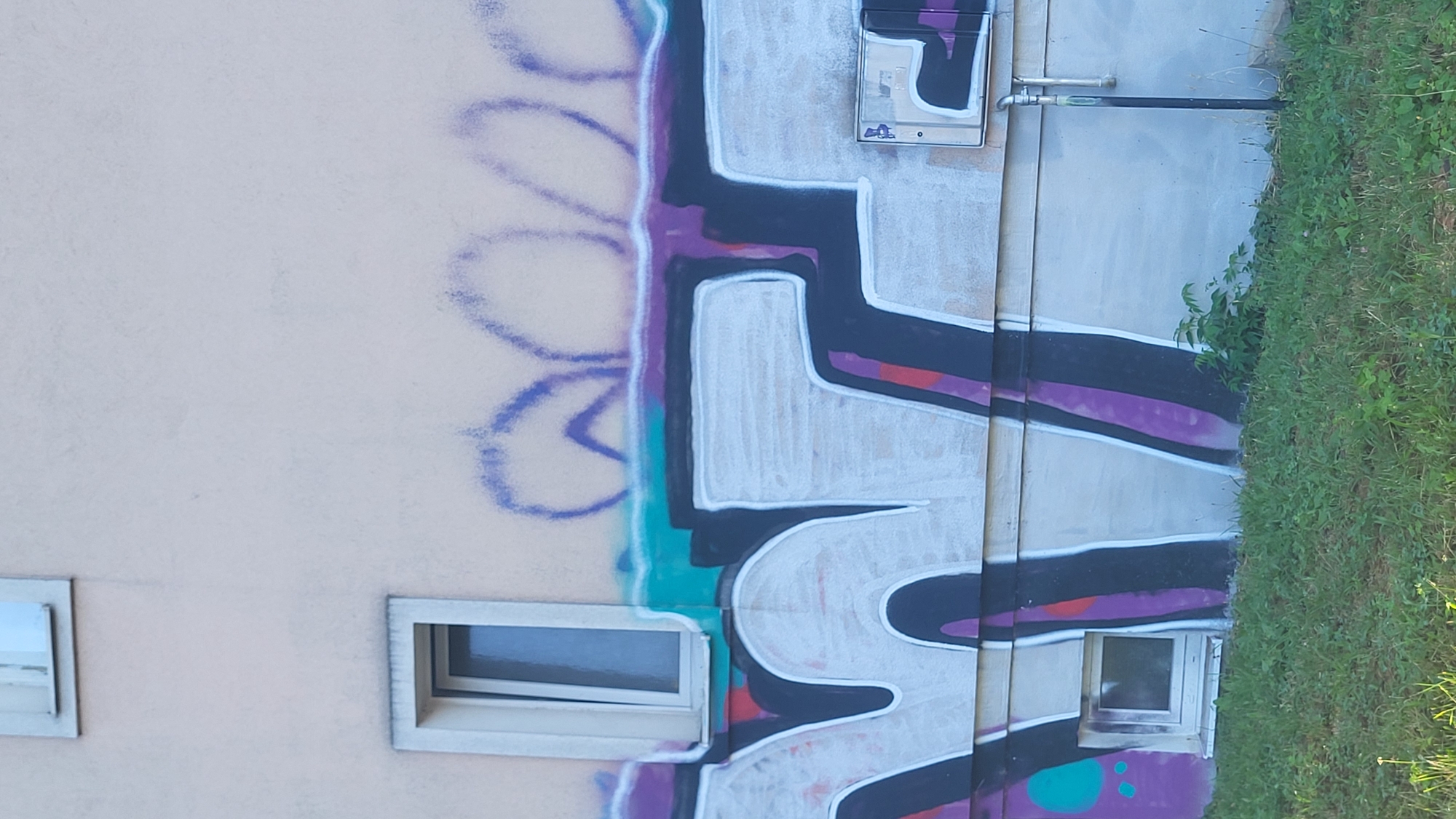 The image shows a section of an exterior wall of a building with graffiti. The graffiti is large and appears to be in a bubble-letter style, primarily in white with dark purple and black outlines. There are also sections of teal and purple spray paint above the main lettering, and some smaller purple and teal sprayed shapes to the left. The wall itself is a light beige color.
There are two windows visible on the wall. The upper window is small and rectangular, with a white frame. The lower window is larger, also rectangular with a white frame, and is partially obscured by the graffiti.
A metal utility box with a reflective surface is mounted on the wall to the right of the graffiti. A pipe runs vertically down the wall from this box.
In the foreground, there is a strip of green grass and some small green plants growing at the base of the wall.
The image appears to have been taken during the daytime, with natural lighting. The perspective is from a low angle, looking up at the wall. There are no people or animals visible in the image. Based on the style of graffiti and the urban setting, this could be a location in the city of Prešov, Slovakia, often found on the exterior walls of apartment buildings or other public structures.