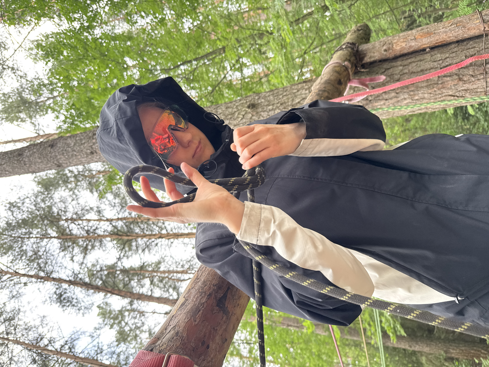 The image shows a person wearing a black hooded jacket and reflective goggles, holding a rope in their hands. The person appears to be in a forest or wooded area, with trees visible in the background. The rope is thick and has a dark and light pattern. There are also other ropes and wooden structures in the background, suggesting an outdoor activity or adventure park. The overall lighting suggests it is daytime, with diffused light filtering through the trees. The location is identified as Chaikovsky, Russia.