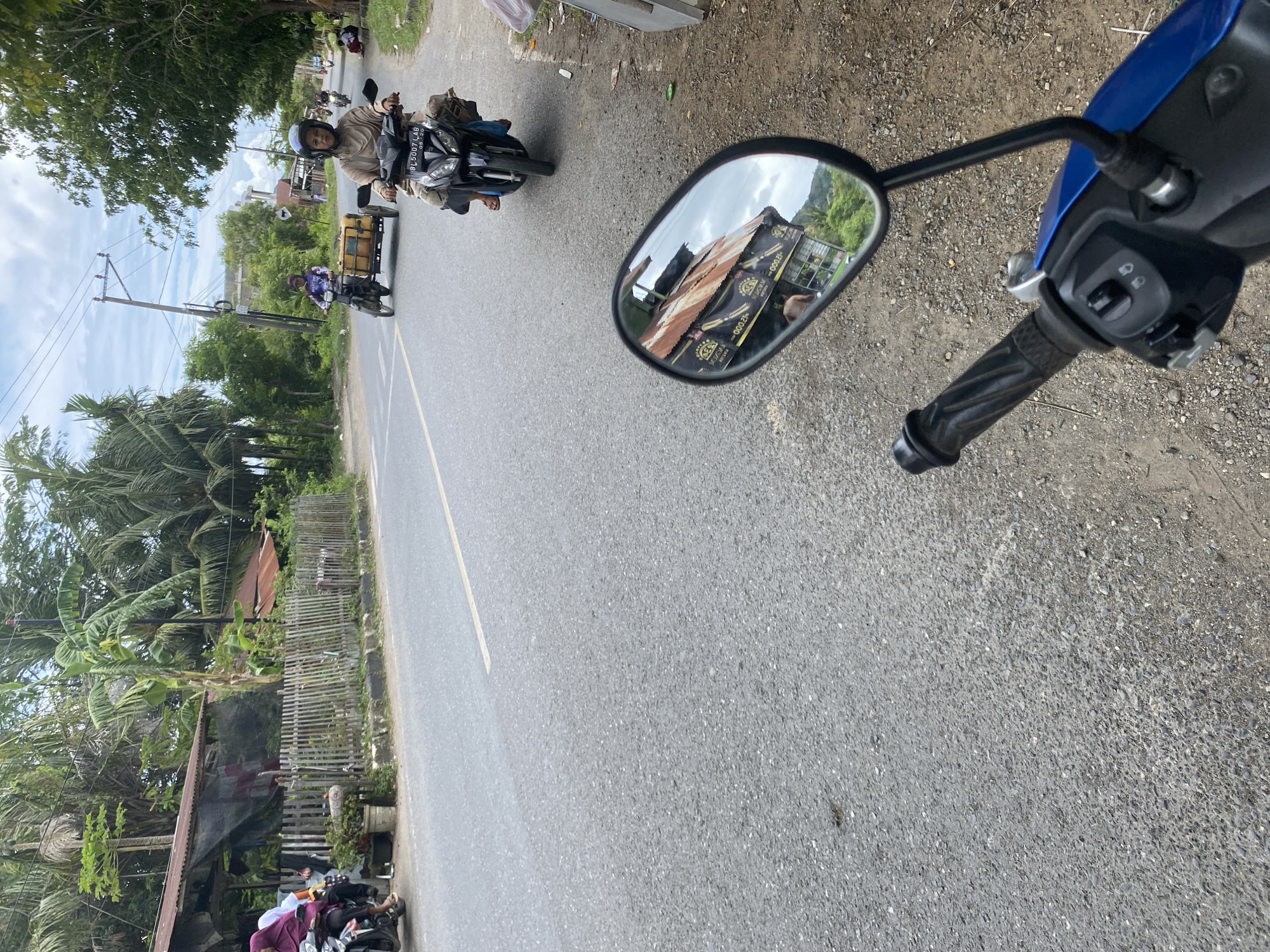 The image captures a street scene in what appears to be a rural or suburban area, possibly in Southeast Asia, given the presence of motorcycles and the vegetation. The perspective is from the point of view of someone on a blue motorcycle, with its handlebar and mirror visible in the foreground. The mirror reflects a building with a rusty metal roof and some signage.
In the midground, a woman wearing a headscarf and loose clothing is riding a black motorcycle. She is wearing a helmet. Further down the road, another individual is riding a motorcycle and pulling a small cart containing yellow containers. This person is also wearing a helmet and is dressed in a similar fashion.
On the left side of the road, there are several structures, including what looks like a small shop or house with a corrugated metal roof and a wooden fence. A person in a purple headscarf is visible on a motorcycle near this structure. The road itself is paved and appears to be a two-lane street with a yellow line marking the center. The environment is lush with tropical trees and vegetation. The sky is partly cloudy. The ground beside the road is unpaved and covered with gravel and dirt. Several other motorcycles are visible in the distance, suggesting moderate traffic.