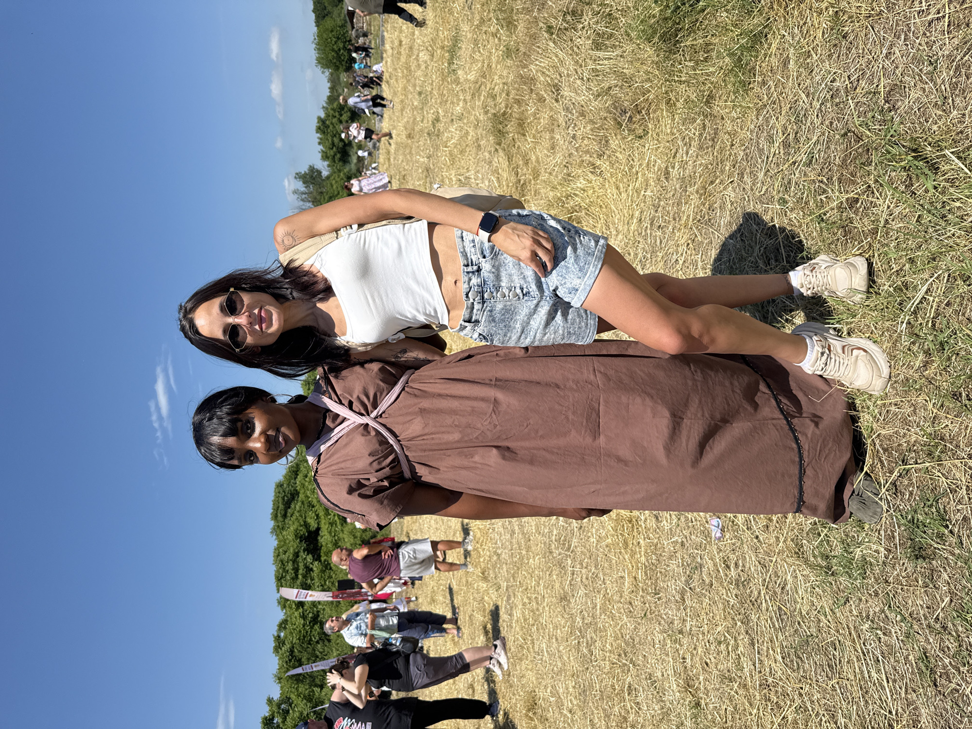 This image depicts two women posing for a photograph outdoors in a dry, grassy field in Gigen, Bulgaria, under a clear blue sky with minimal scattered white clouds.
The woman on the left, with dark skin and dark hair styled with bangs, is wearing a long, loose, mocha-brown dress. The dress features a pale pink or light-colored criss-cross strap design across the chest, creating a gathered effect on the bodice. A thin black beaded trim runs along the lower portion of the dress. She wears large gold-toned hoop earrings and dark, flat shoes that are partially obscured by the dress hem. She is looking directly at the camera with a slight smile.
The woman on the right, with lighter skin and long dark hair, is wearing dark hexagonal sunglasses. She is dressed in a white sleeveless cropped tank top, revealing her midriff. Her lower attire consists of light blue acid-wash denim shorts with a button-fly closure. On her left wrist, she wears a red-banded smartwatch. A tattoo of a sun is visible on her left upper arm. She has a tan or beige shoulder bag slung across her body. Her footwear consists of chunky, off-white or beige athletic sneakers with thick soles. She is standing with her left leg slightly bent and her left hand resting on her hip/shorts, looking directly at the camera with a slight smile.
Both women are standing side-by-side, appearing to be in conversation or posing together. The ground beneath them is covered in dry, golden-brown grass and some patches of green, indicating a warm, sunny environment.
In the mid-ground and background, numerous other individuals and small groups are visible, suggesting an outdoor event or gathering. To the left, a man in a black t-shirt and cap, another man in a light blue collared shirt, and a man in a burgundy tank top and white shorts are present. White banners or flags on poles are also visible. A line of green trees forms the distant horizon, separating the field from the sky.