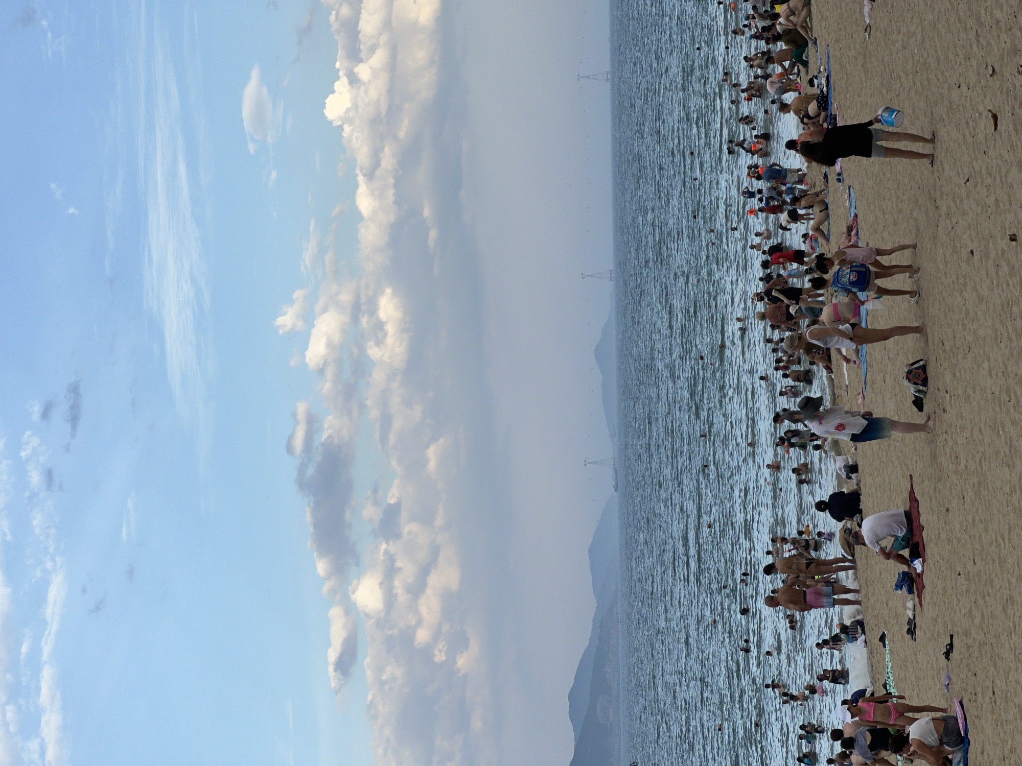 The image depicts a crowded beach scene in Nha Trang, Vietnam. In the foreground, people are relaxing on the sandy shore, some sitting on towels and others standing. A few individuals are wading into the shallow water at the edge of the sea. Further out, a large number of people are swimming and playing in the ocean. The sea is a deep blue and moderately wavy, extending towards a hazy horizon where distant mountains are visible. The sky above is a mix of pale blue with scattered white and grey clouds. The beach appears to be a popular recreational spot, with many people enjoying the water and sun.