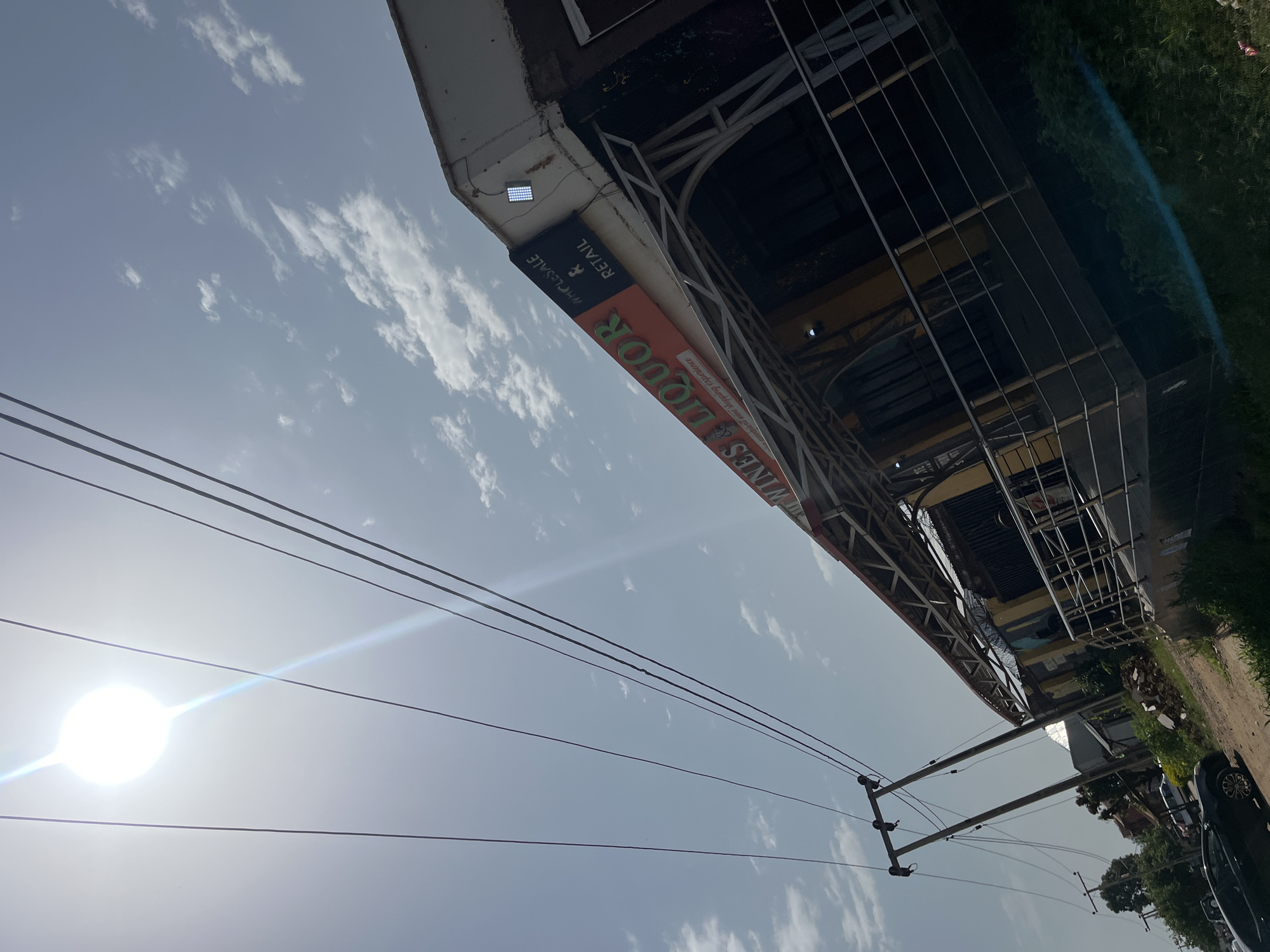 This media file captures an outdoor scene in Bukuru, Nigeria, during what appears to be daytime. The image is oriented sideways, showing a bright sky with scattered white clouds and a strong sun casting a lens flare. In the foreground, several power lines are visible against the sky.
A building with a metallic structure and a light-colored overhang dominates the upper portion of the frame. A sign on the building reads "LIQUOR" in large, bold letters, with smaller text below it that is partially obscured. Another section of the sign reads "RETAIL" and "WHOLESALE" with a symbol of a woman. The building appears to be a commercial establishment.
To the right of the building, a grassy area and a dirt path are visible. Some vegetation and what looks like construction debris are present near the building's base. A dark-colored car is parked on the right side of the frame, partially obscuring the grassy area and path. Power poles with electrical lines are also visible in the lower half of the image. The overall impression is of a street view in a developing area, possibly a commercial district. The bright sunlight suggests it is likely midday.