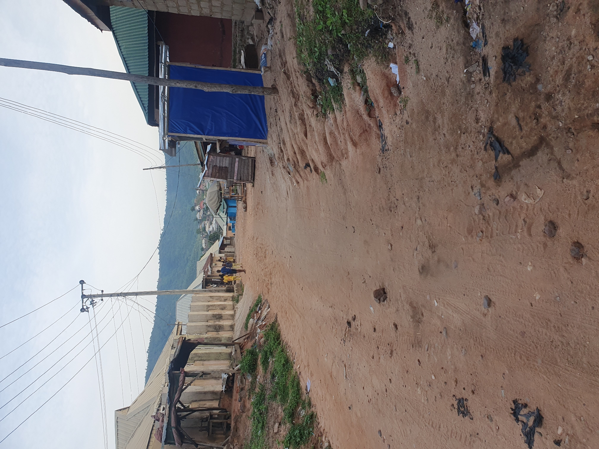 A dirt road winds through a community in Lokoja, Nigeria, flanked by buildings and natural elements. On the left, a row of structures with corrugated metal roofs and rough concrete walls line the road. Some of these buildings appear to be shops or workshops, with open fronts and weathered materials. A wooden structure with a makeshift roof stands out, showing signs of disrepair. Further down the road, a group of three young males, dressed in casual clothing, are walking away from the camera.
On the right side of the road, a blue tarp is draped over a wooden frame, possibly serving as a temporary shelter or enclosure. Adjacent to this, a building with a green corrugated metal roof and brick walls is visible. The terrain on this side of the road slopes upwards, with exposed reddish-brown soil and sparse vegetation. Small stones and debris are scattered across the road and the roadside.
In the background, a verdant, tree-covered hill rises, with a cluster of houses visible on its slopes and at its base. Power lines crisscross the sky, supported by a utility pole with multiple wires. The sky is overcast, suggesting a diffused light. The overall scene depicts a somewhat unkempt, but inhabited, rural or semi-urban environment, typical of parts of Lokoja.