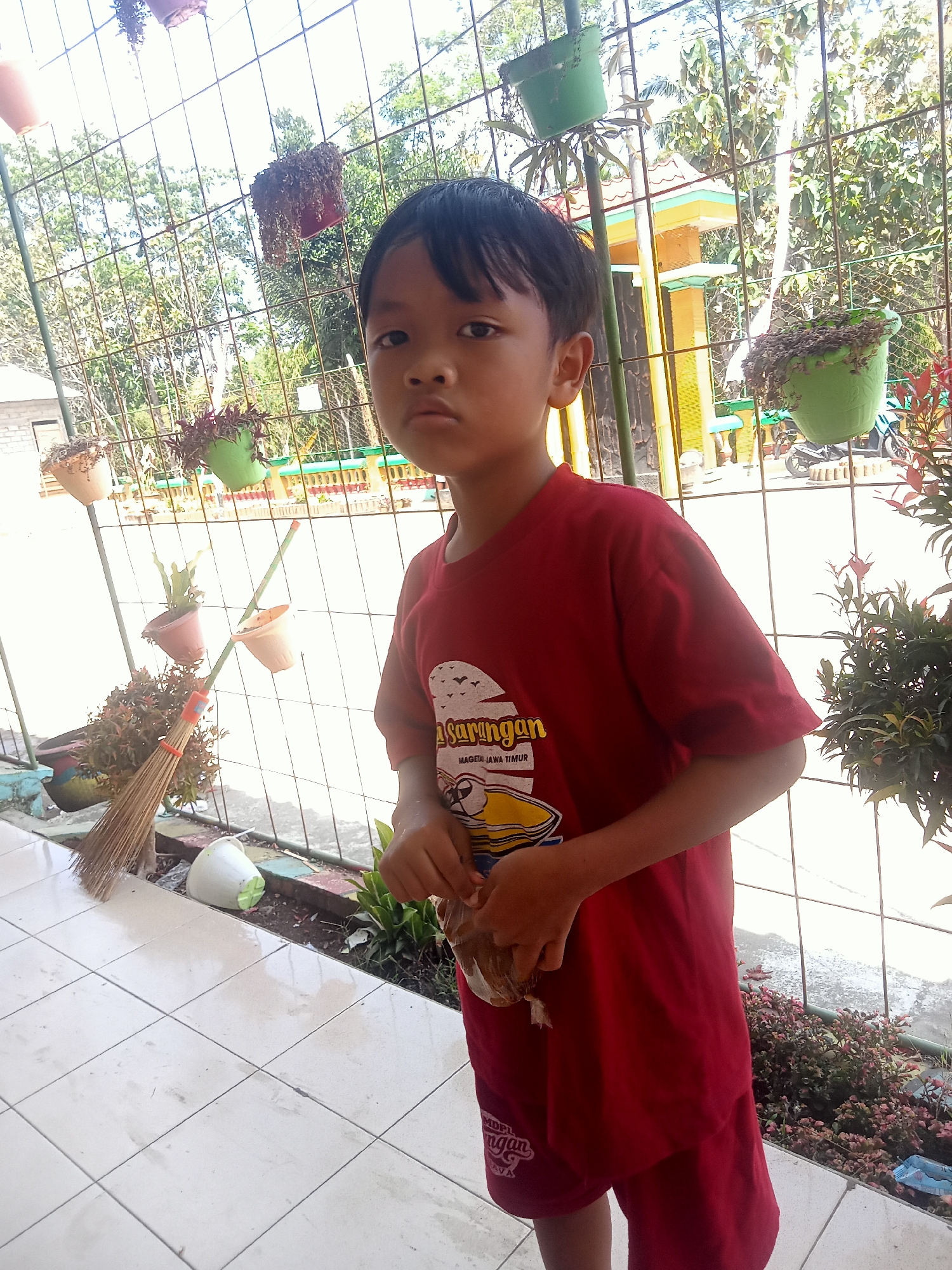 A young Indonesian boy stands in front of a metal fence. He is wearing a red t-shirt with a graphic design and red shorts. The boy is holding a small, wrapped object in his hands. Behind him, several potted plants hang from the fence and are placed on the ground. A broom leans against the fence to his left. The background reveals trees and a building with a yellow and red roof. The scene appears to be outdoors in a residential area of Pule, Indonesia.