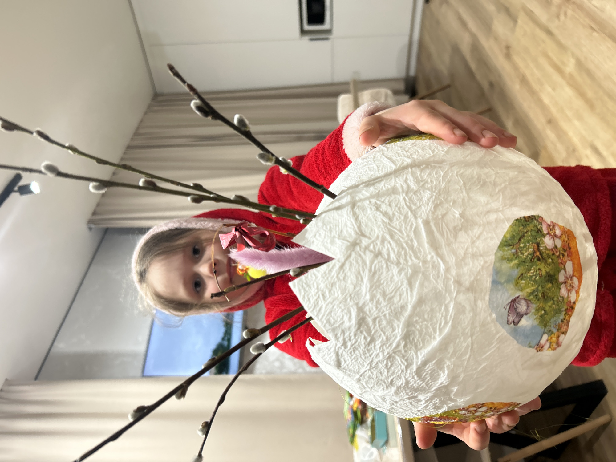 A young girl with blond hair and glasses, wearing a red robe, is holding a large white decorated egg with pussy willow branches sticking out of the top. The egg has a butterfly sticker on it. The scene is indoors, in a modern-looking apartment. The location is likely Chaikovsky, Russia.